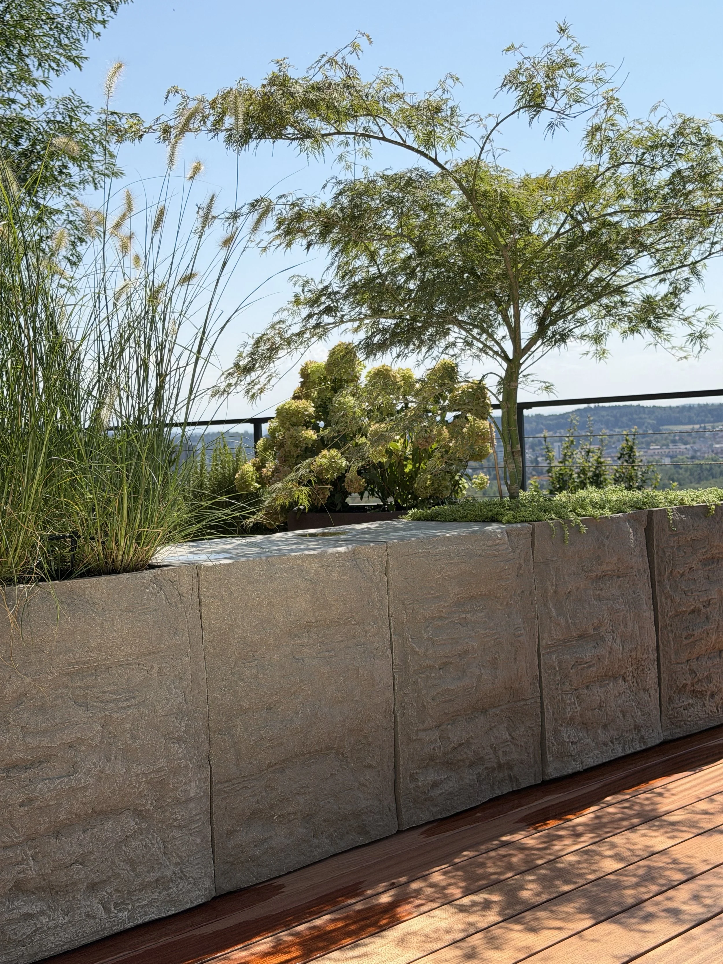 Ein Balkon mit einer Steinmauer, auf der viele Pflanzen wachsen, darunter ein Baum, Gräser und Blumen, mit Blick auf eine Stadt im Hintergrund bei sonnigem Wetter.