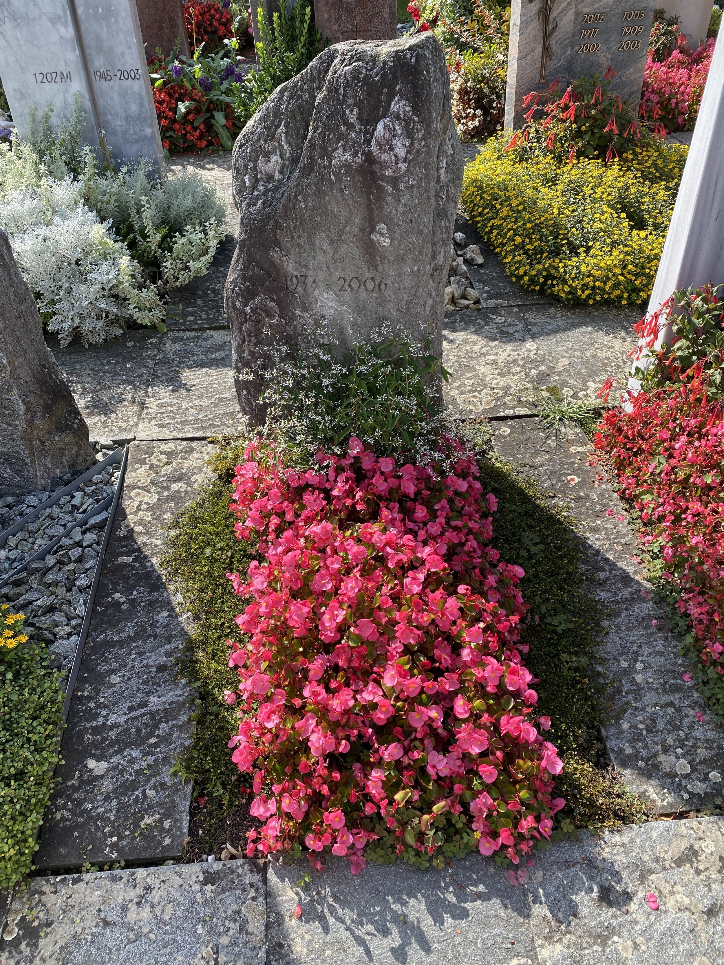 Grabstein mit Jahreszahlen in einem Friedhof, umgeben von buntem Blumen und Pflanzen.