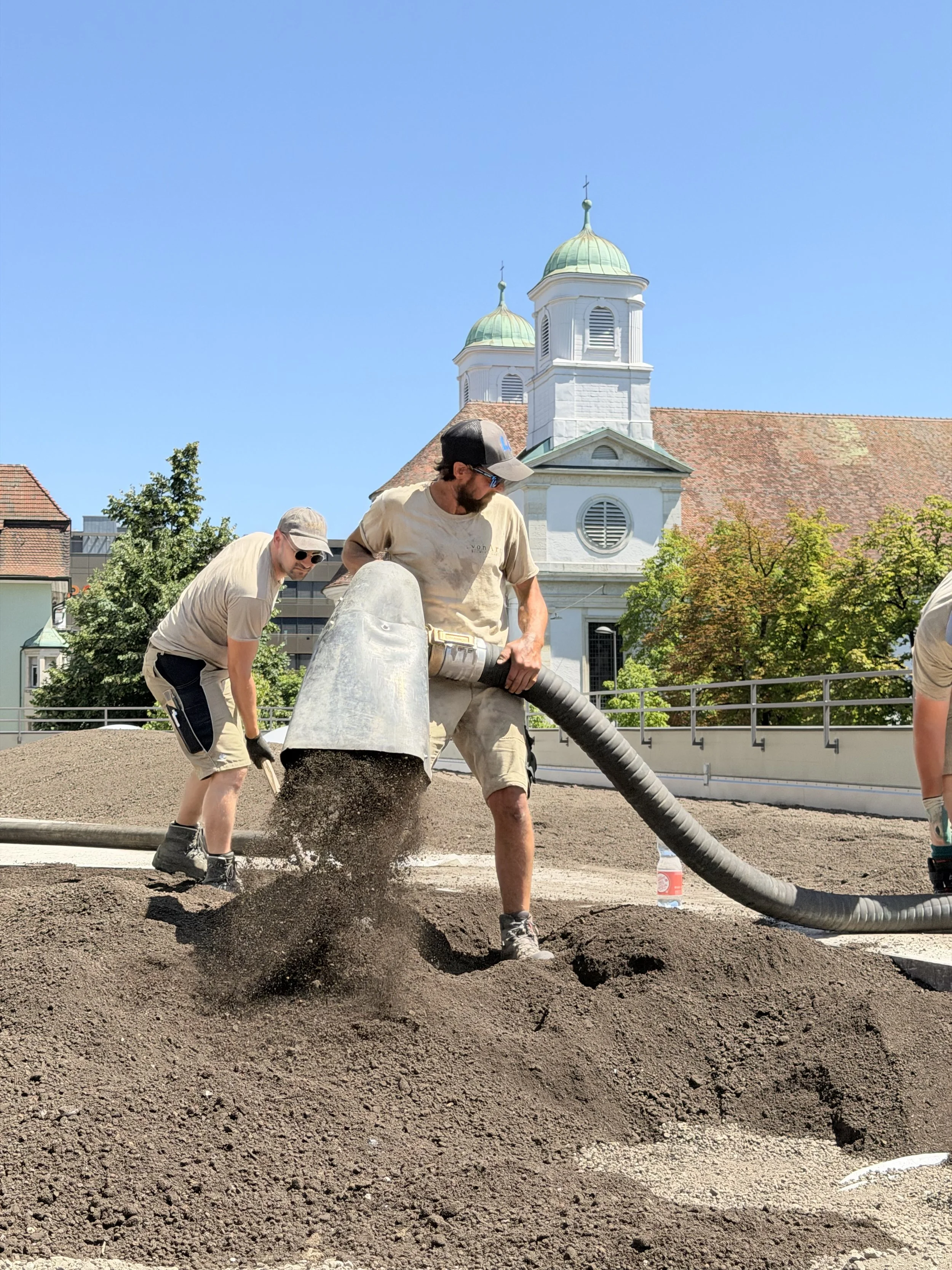 Arbeitsleute verteilen Erde auf einer Baustelle, im Hintergrund eine Kirche mit zwei Türmen und grünen Kuppeln, im Vordergrund sind zwei Männer bei der Arbeit mit einem großen Schlauch.