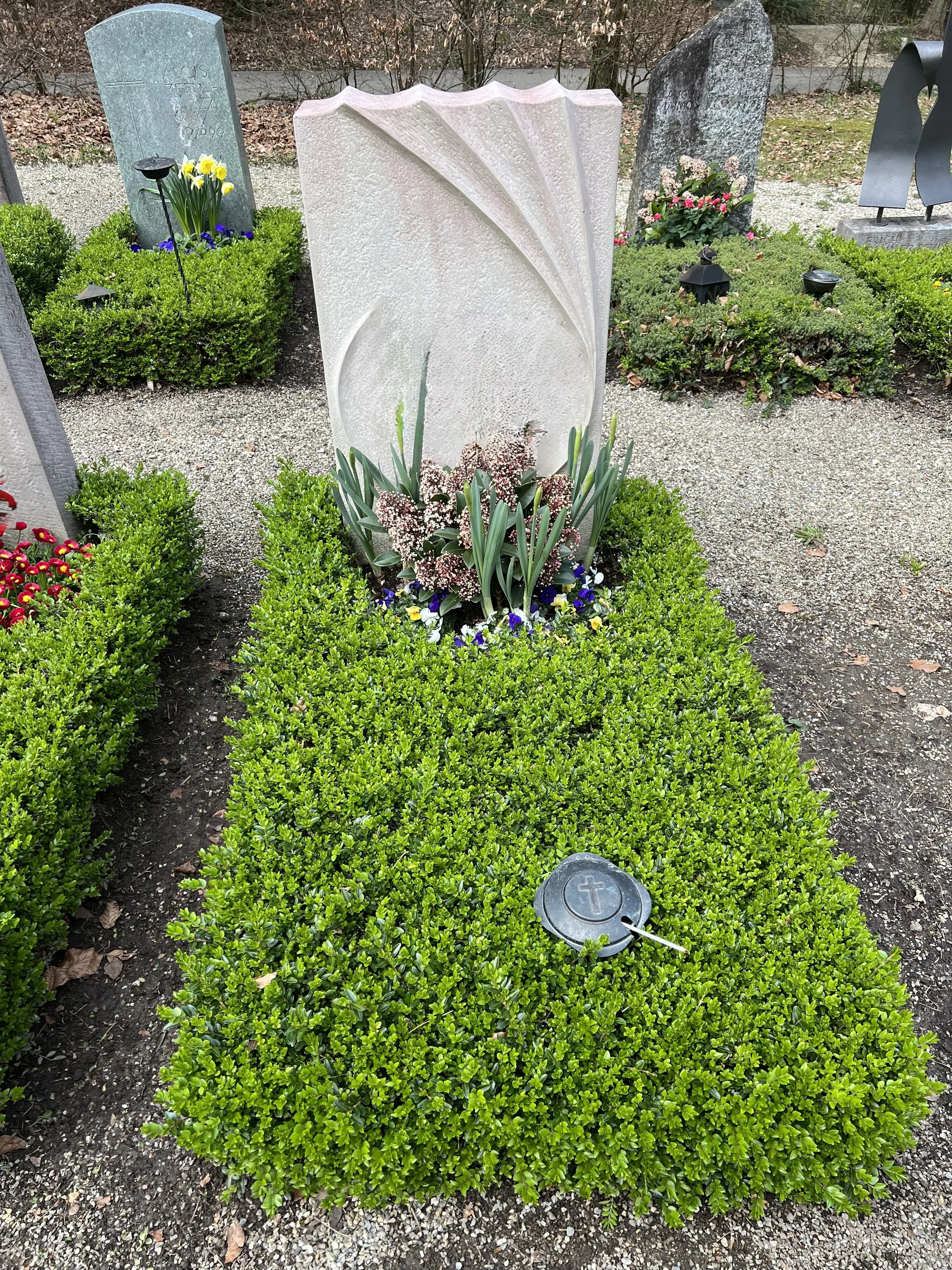 Grüner Friedhof mit Gräbern, Blumen, Kerzen und einem Grabstein. Im Vordergrund ein Grab mit einer Glocke und einem Kreuz.