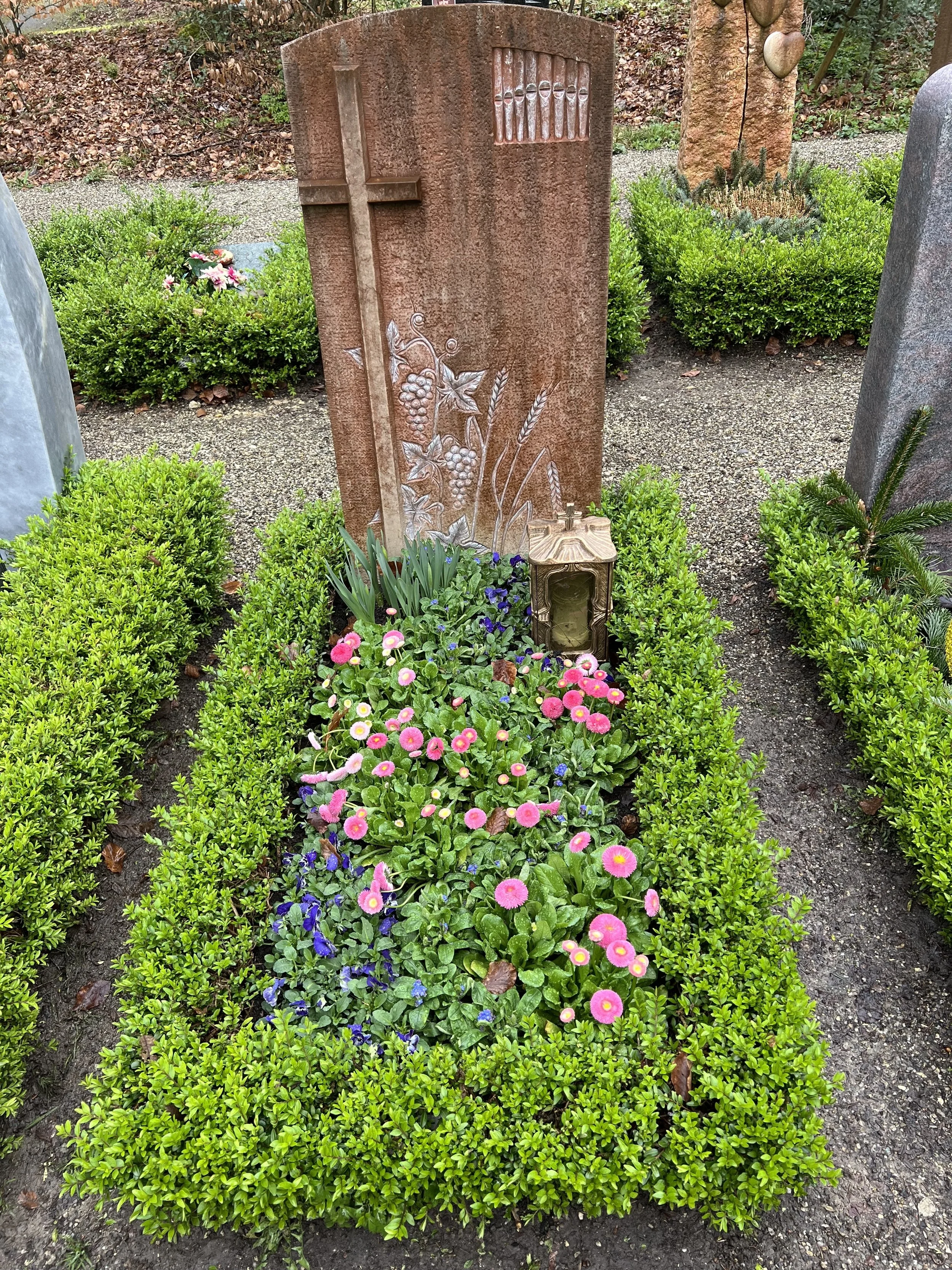 Ein Grabstein auf einem Friedhof, umgeben von bunten Blumen und grünen Büschen. Der Stein ist verziert mit einem Pfahl, Weinreben und Getreideähren.
