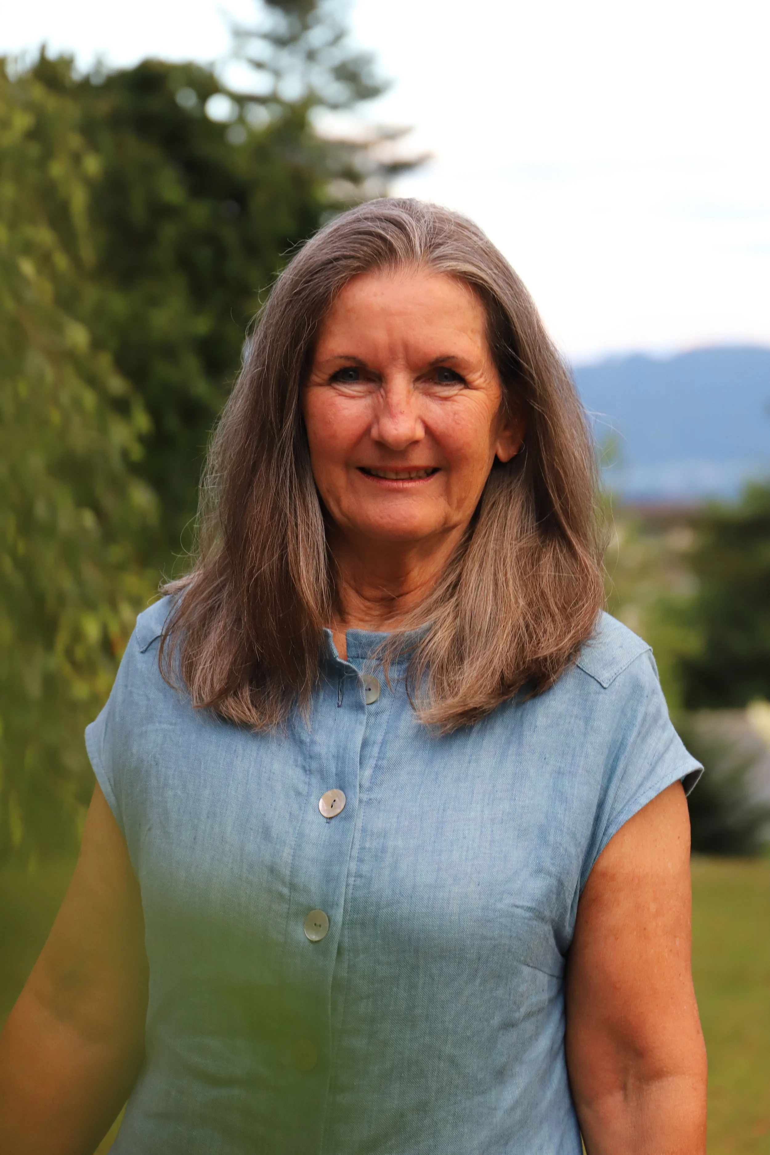 Ein älterer Frau mit langen braunen Haaren, die lächelnd im Freien steht, mit grüner Vegetation im Hintergrund.