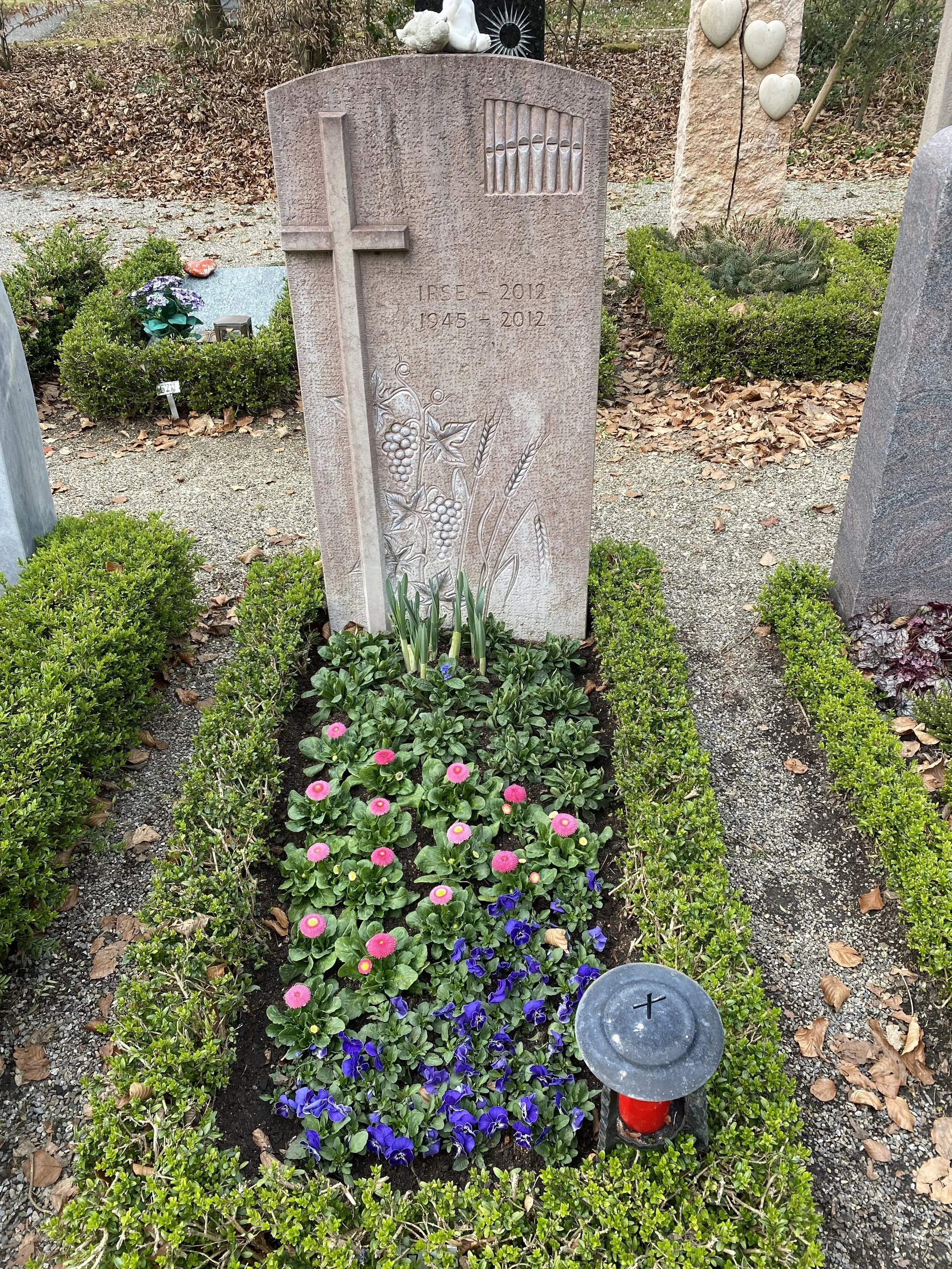 Grabstein mit Blumen und Kerze, umliegende Büsche und leere Flächen, im Hintergrund Bäume und Laub.