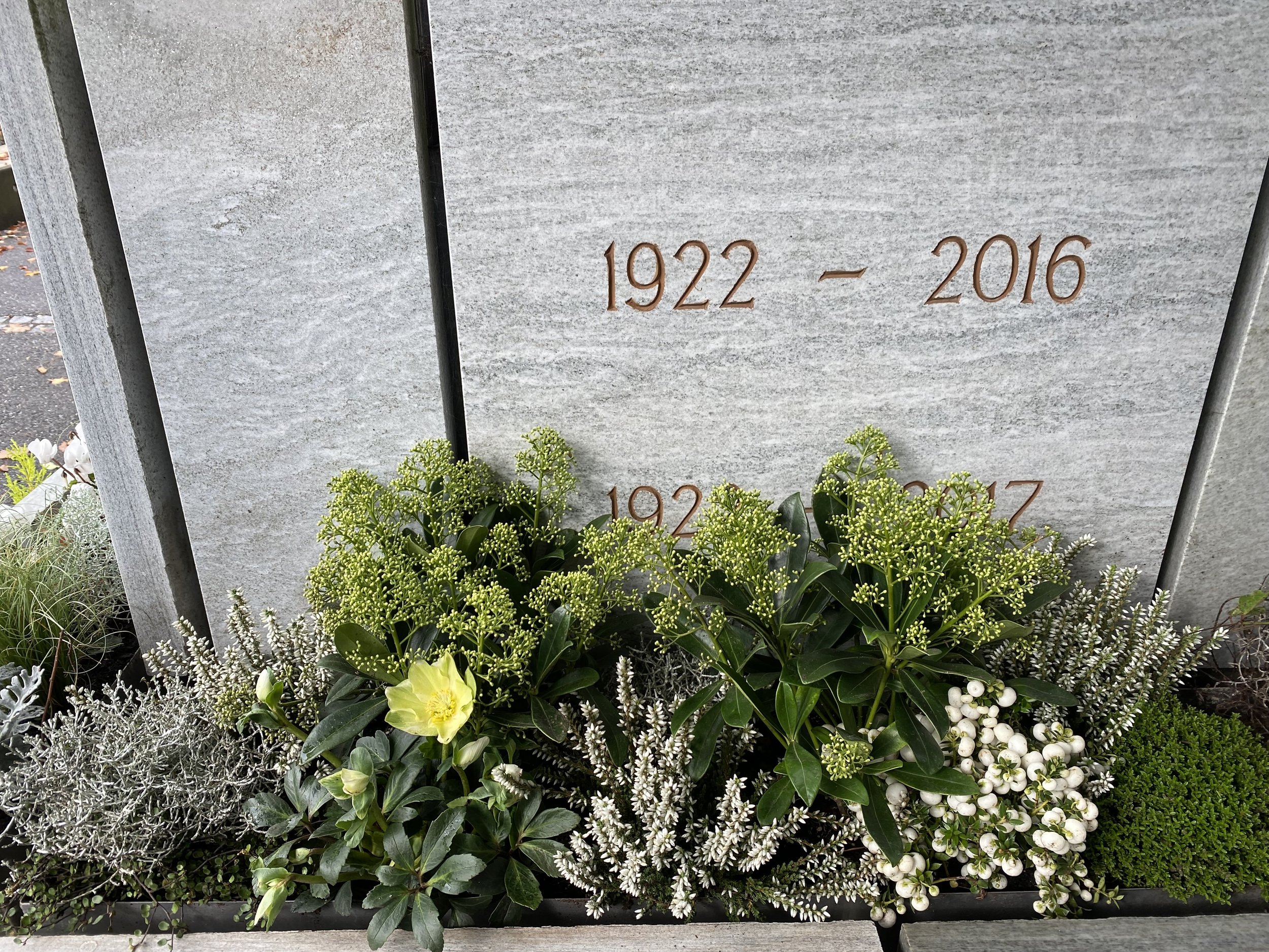 Gedenktafel mit den Jahren 1922–2016 und grüne und weiße Blumen darunter.