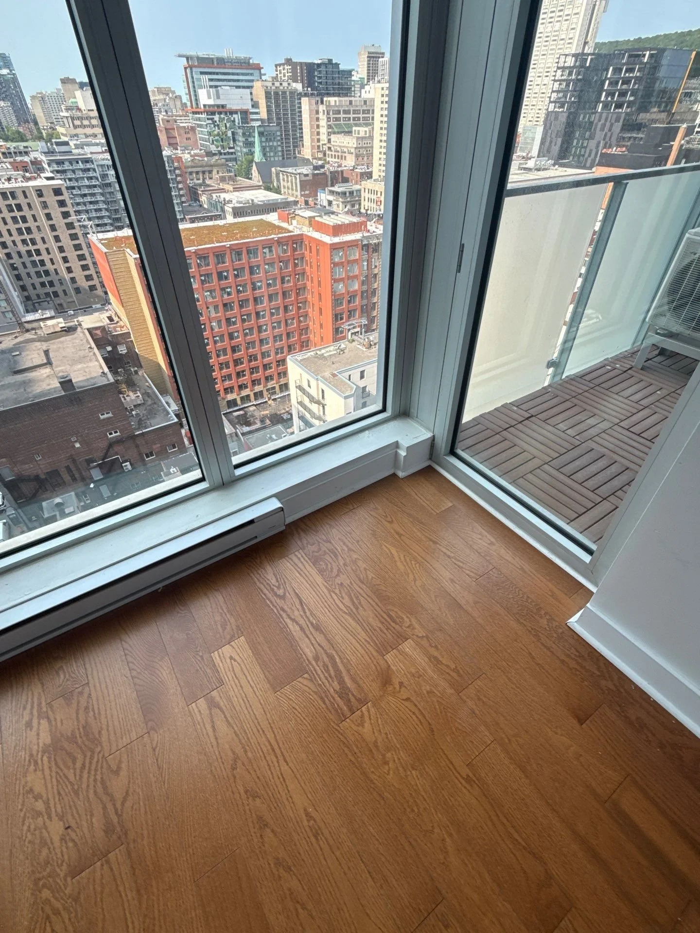 Interior of a high-rise apartment with a wooden floor, large glass windows, and a balcony showing city skyscrapers.