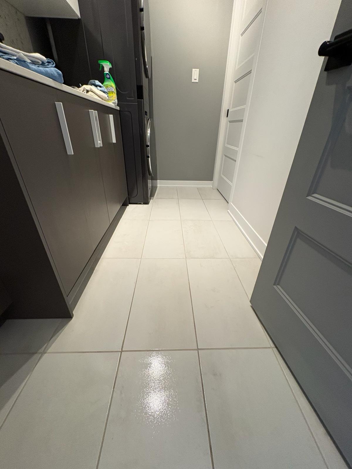 A laundry room with a tiled floor, gray cabinets, a black washing machine, a white door, and cleaning supplies on the counter.