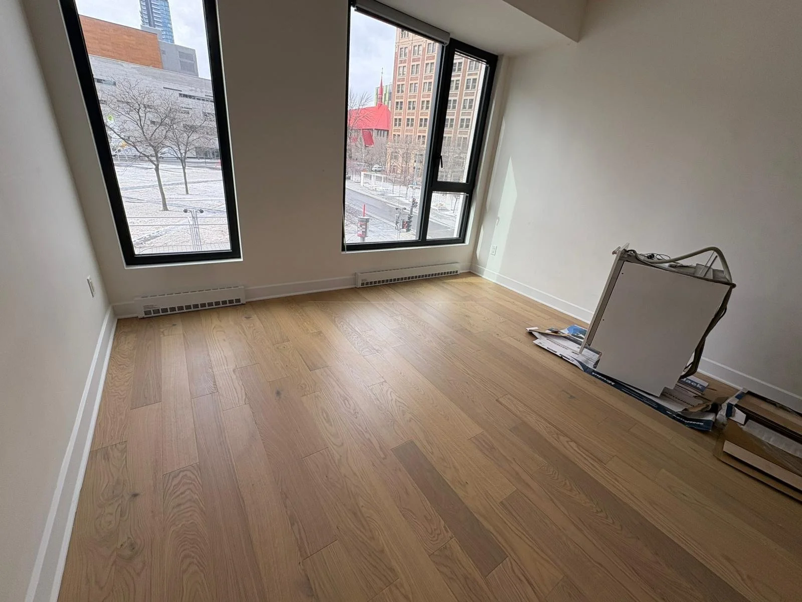 Empty room with large windows, light wood flooring, and some unpacked boxes and a foldable chair in the corner, indicating recent moving or renovation.