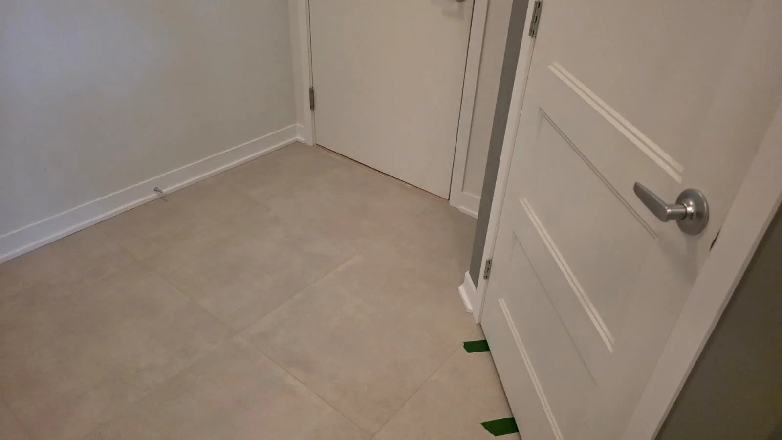 Interior corner of a room with beige tiled floor, white baseboard, and two white doors, one with a silver handle.