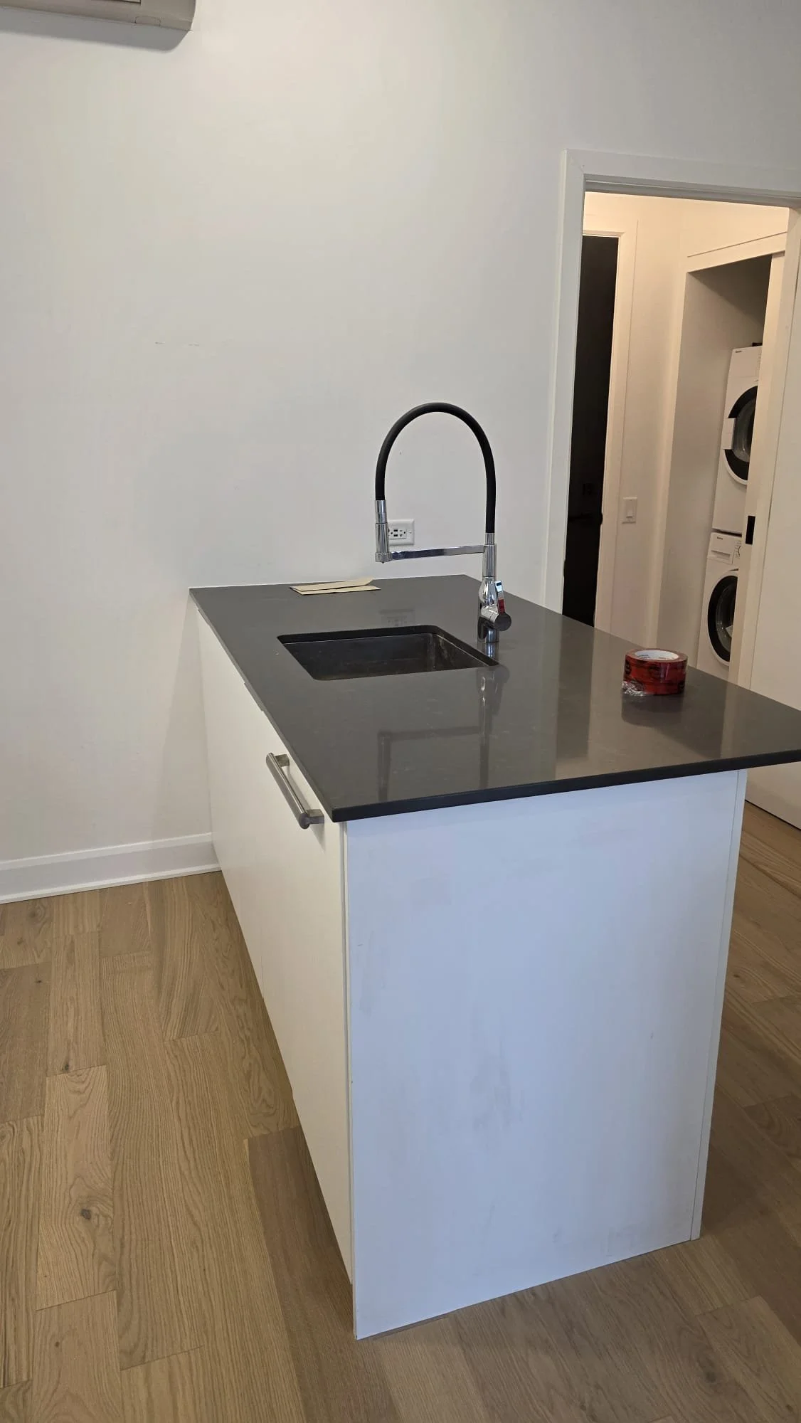 A small kitchen island with a dark gray countertop and white base, featuring a black faucet and sink. On the counter is a roll of red tape, and the background shows a laundry area with a stacked washer and dryer.
