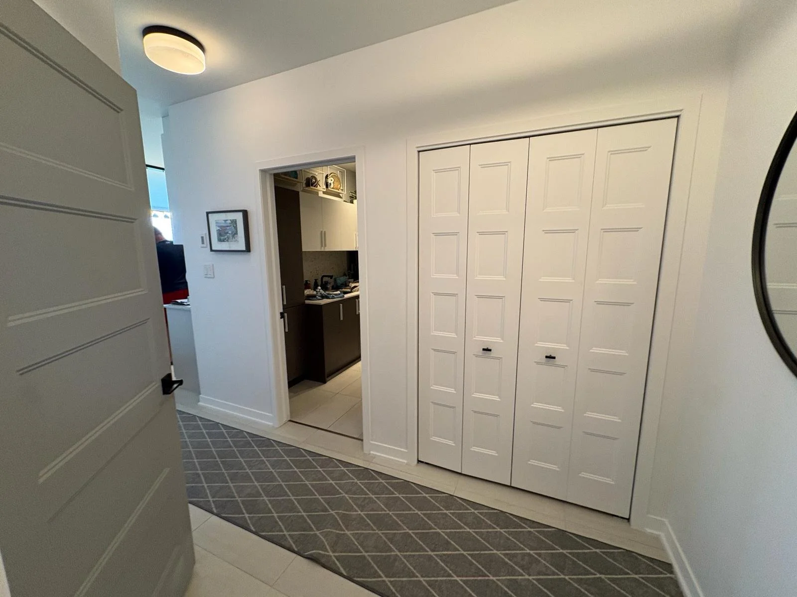 Interior view of a room with a white wall, a closet with folding doors, a doorway leading to a kitchenette, and a patterned rug on light-colored tile flooring.