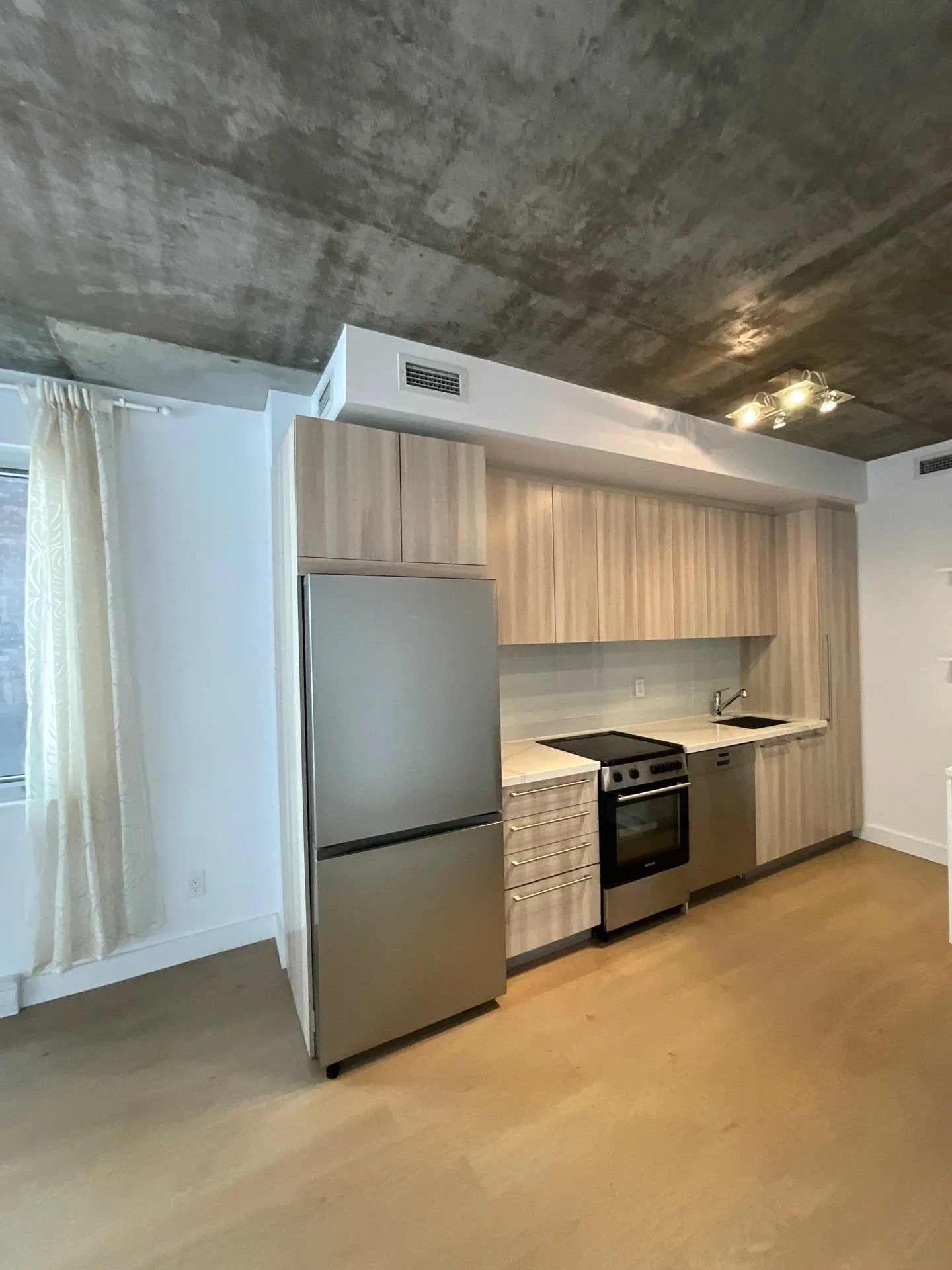 Modern kitchen with light wood cabinets, stainless steel refrigerator, stove, and small sink, with beige flooring and a concrete ceiling.