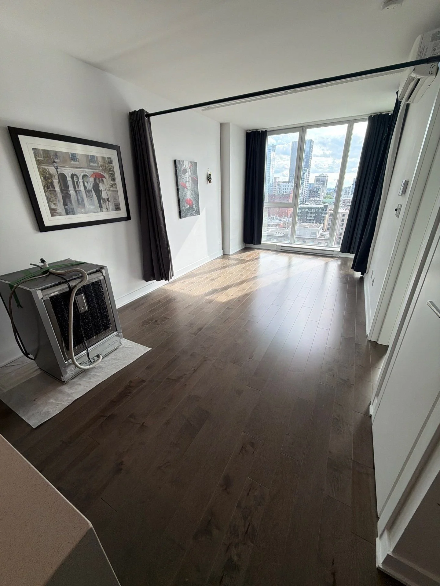 A room with wooden flooring, large window with cityscape view, and wall art. There is a piece of equipment on a mat in the corner.