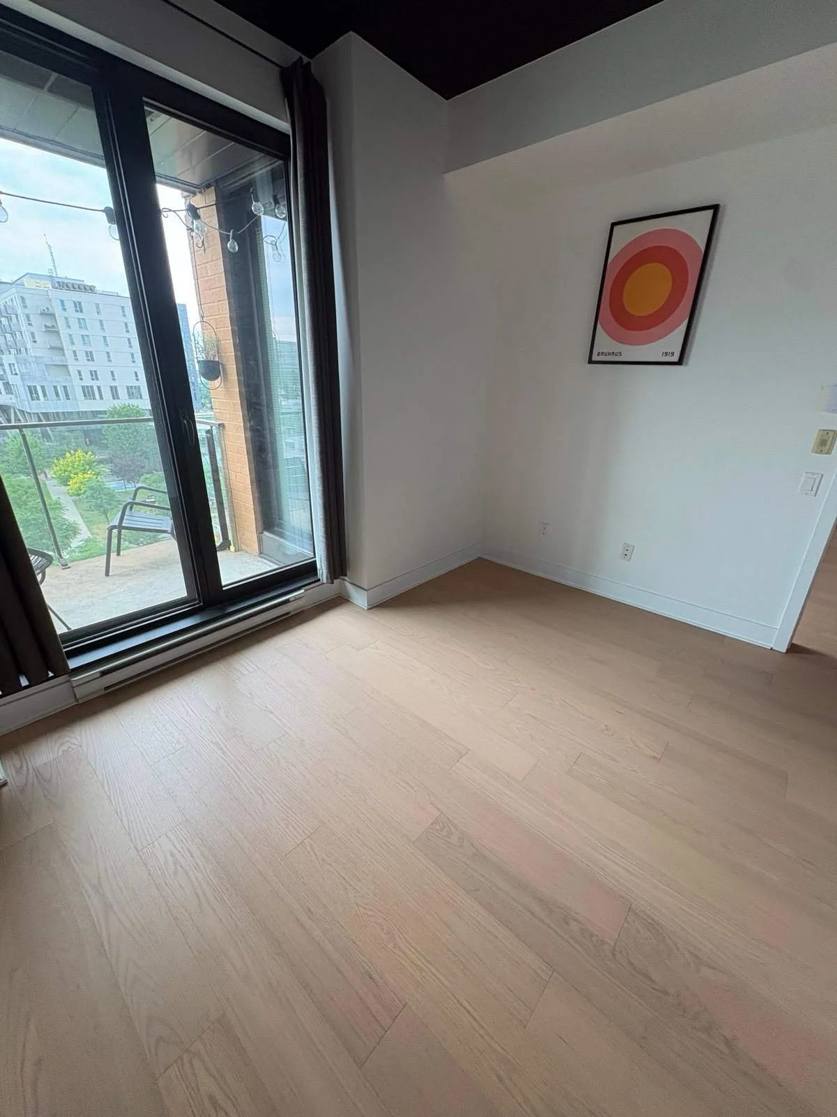 Empty living room with wooden flooring, a large sliding glass door leading to a balcony, and a framed abstract artwork on the wall.