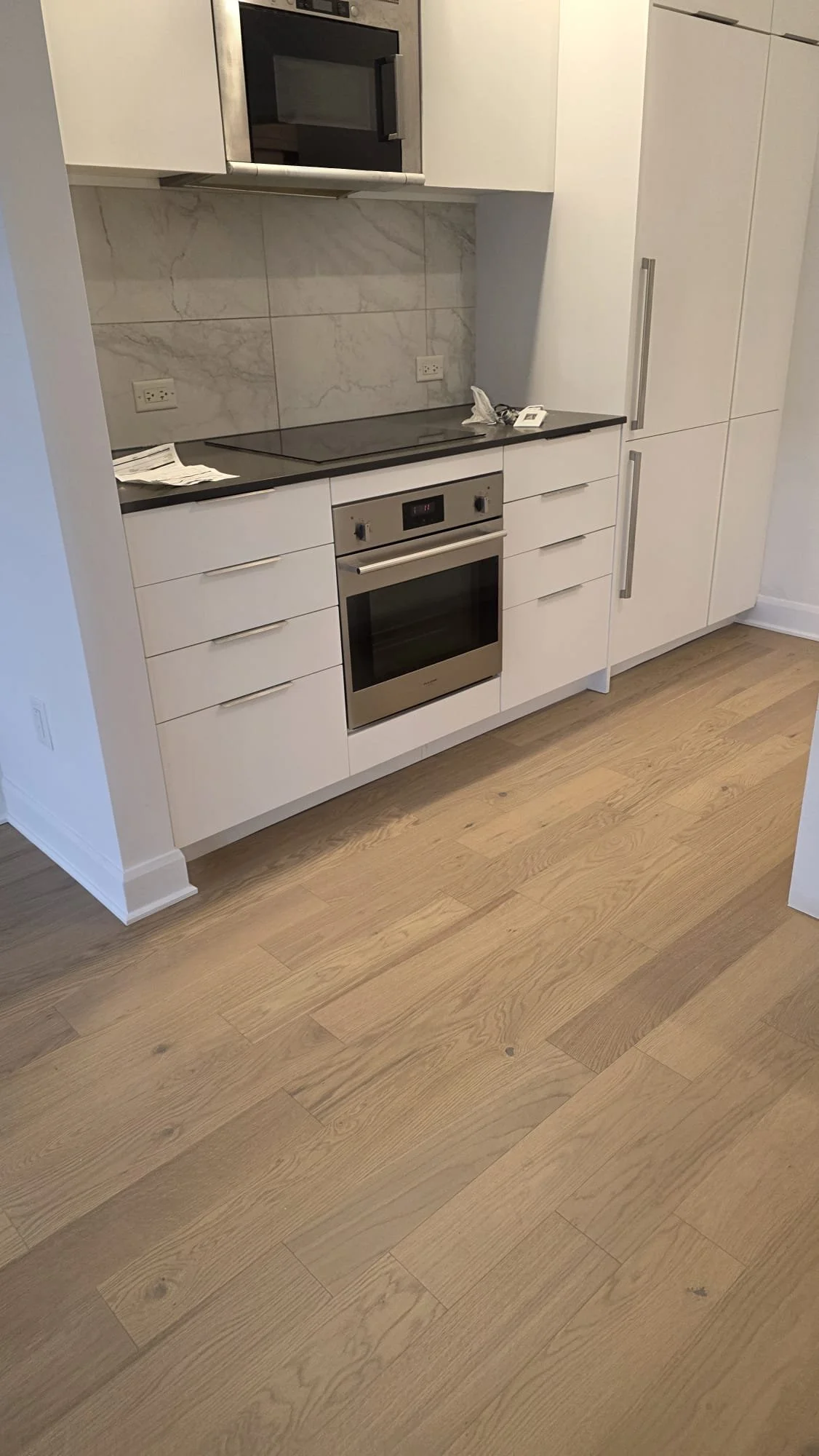 Modern white kitchen with black countertop, stainless steel oven, microwave, and integrated refrigerator, light wooden flooring, marble backsplash, and some scattered items on the counter.