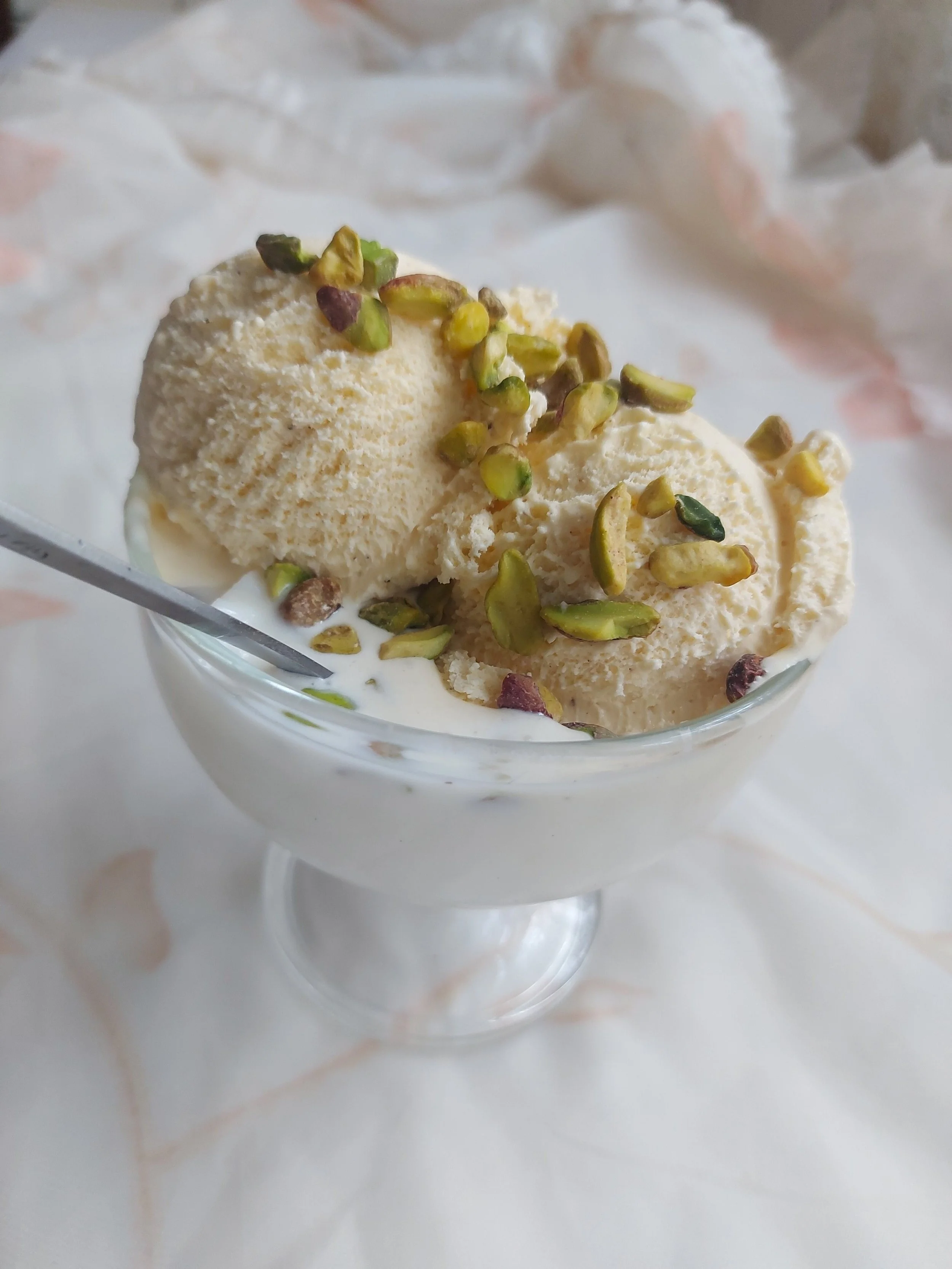 Two scoops of vanilla ice cream topped with chopped pistachio nuts in a glass dessert dish with a spoon.
