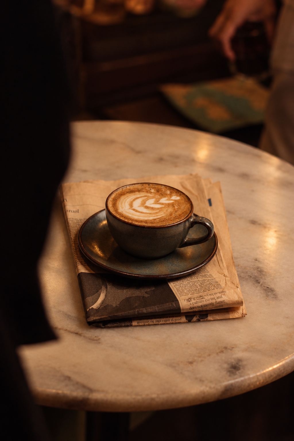 A cup of latte with latte art on top placed on a black saucer, resting on a folded newspaper on a marble table in a cozy café setting.