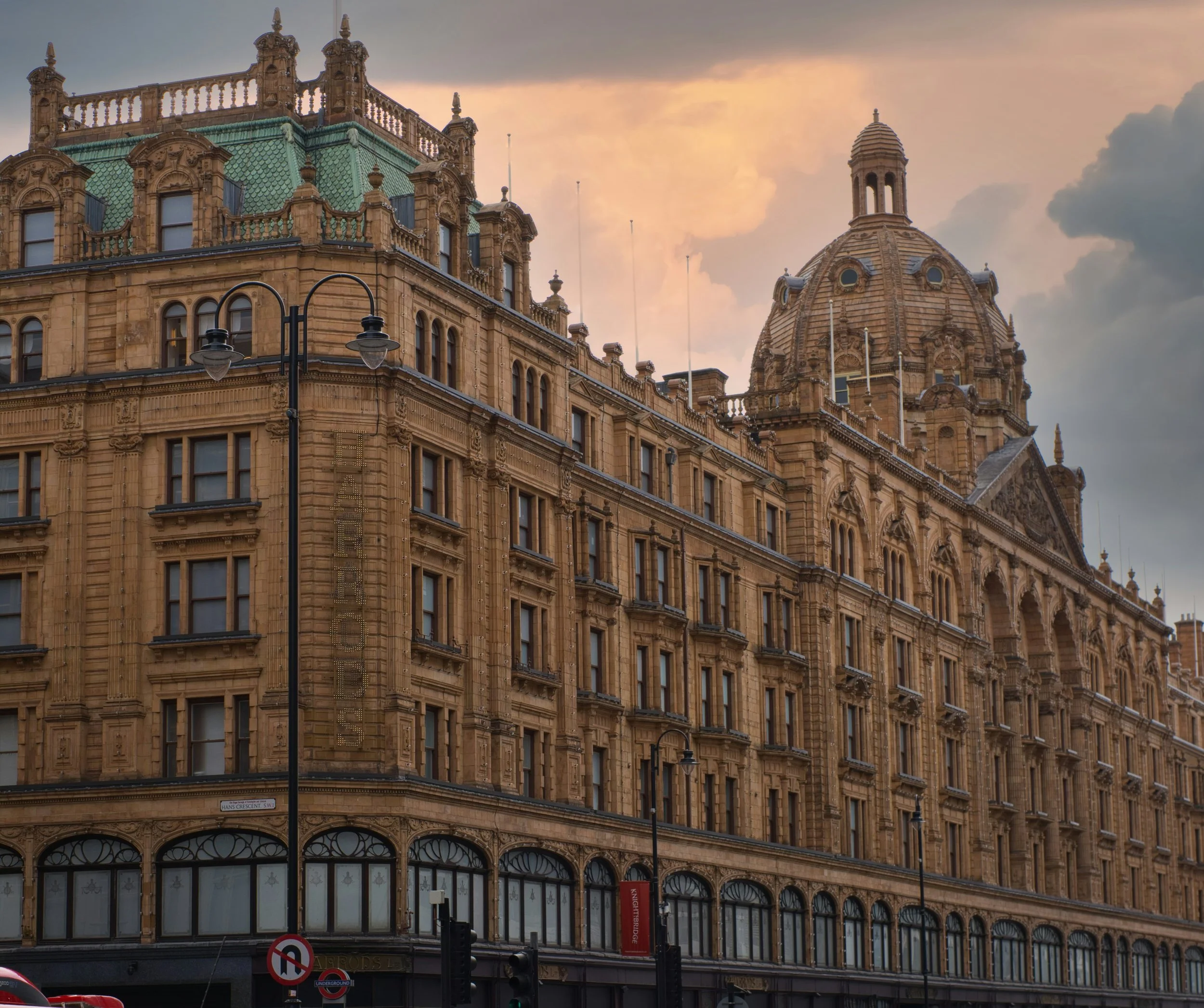 Historic stone building with ornate rooftops and a domed tower, city street with streetlights and signs, sunset sky with clouds.