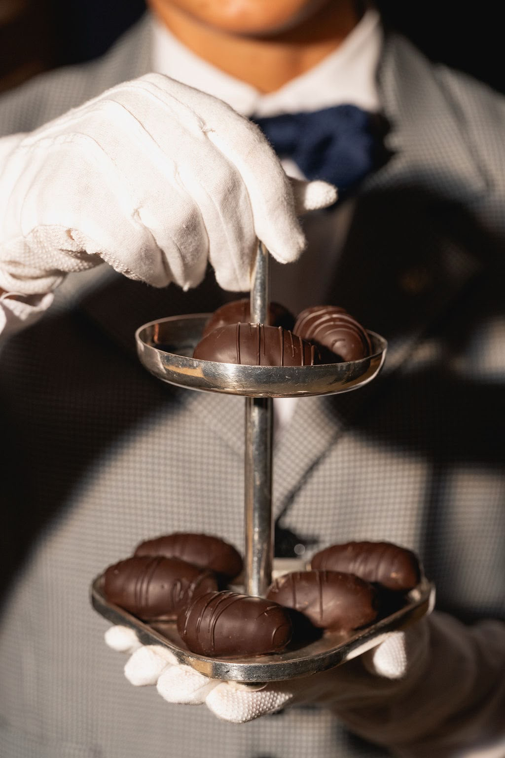 Person wearing white gloves, a checkered suit, and a blue bow tie, serving chocolate truffles on a tiered platter.
