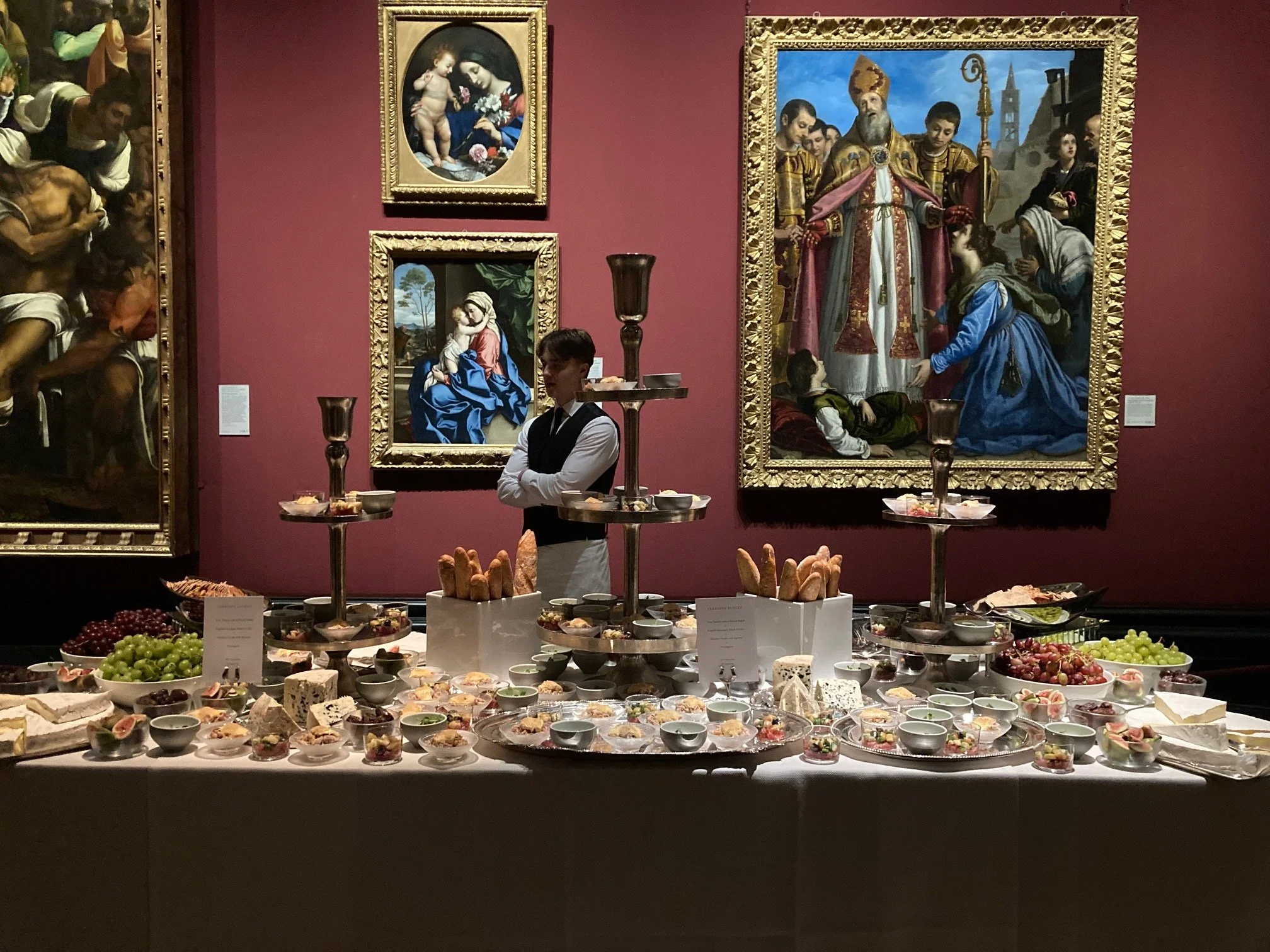A display of assorted cheeses, fruits, and appetizers set on a table in front of classic religious artwork paintings in an art gallery.