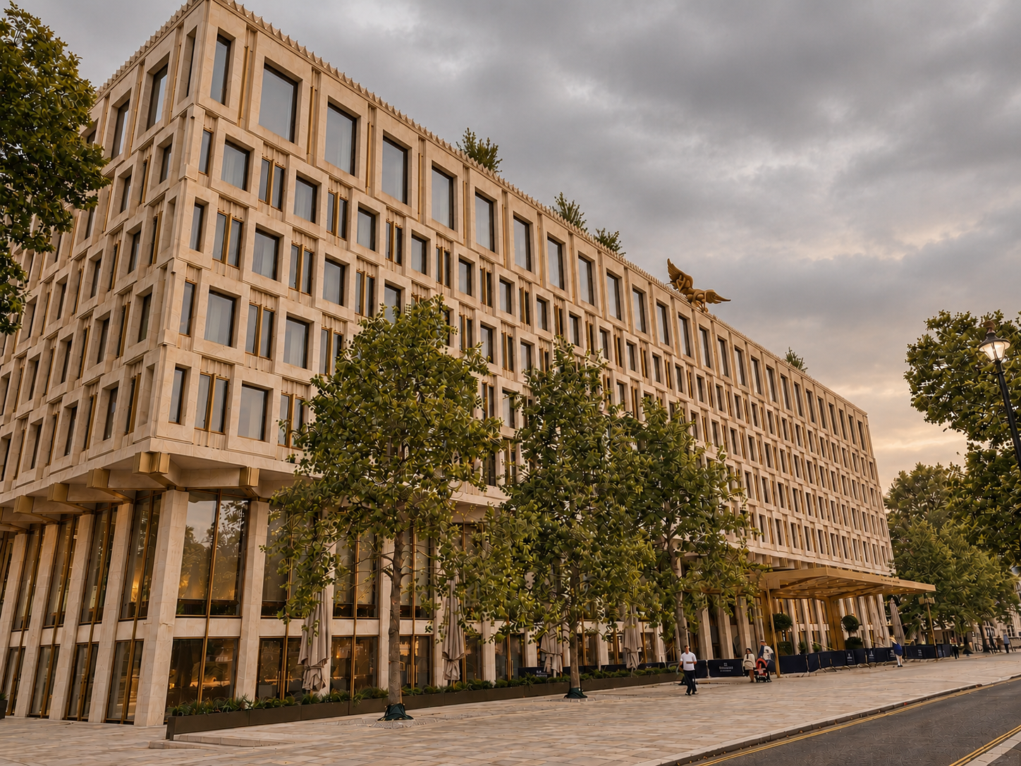A large, modern multi-story building with numerous windows, trees in front, and a paved walkway with a few people walking near the entrance. The sky is overcast.