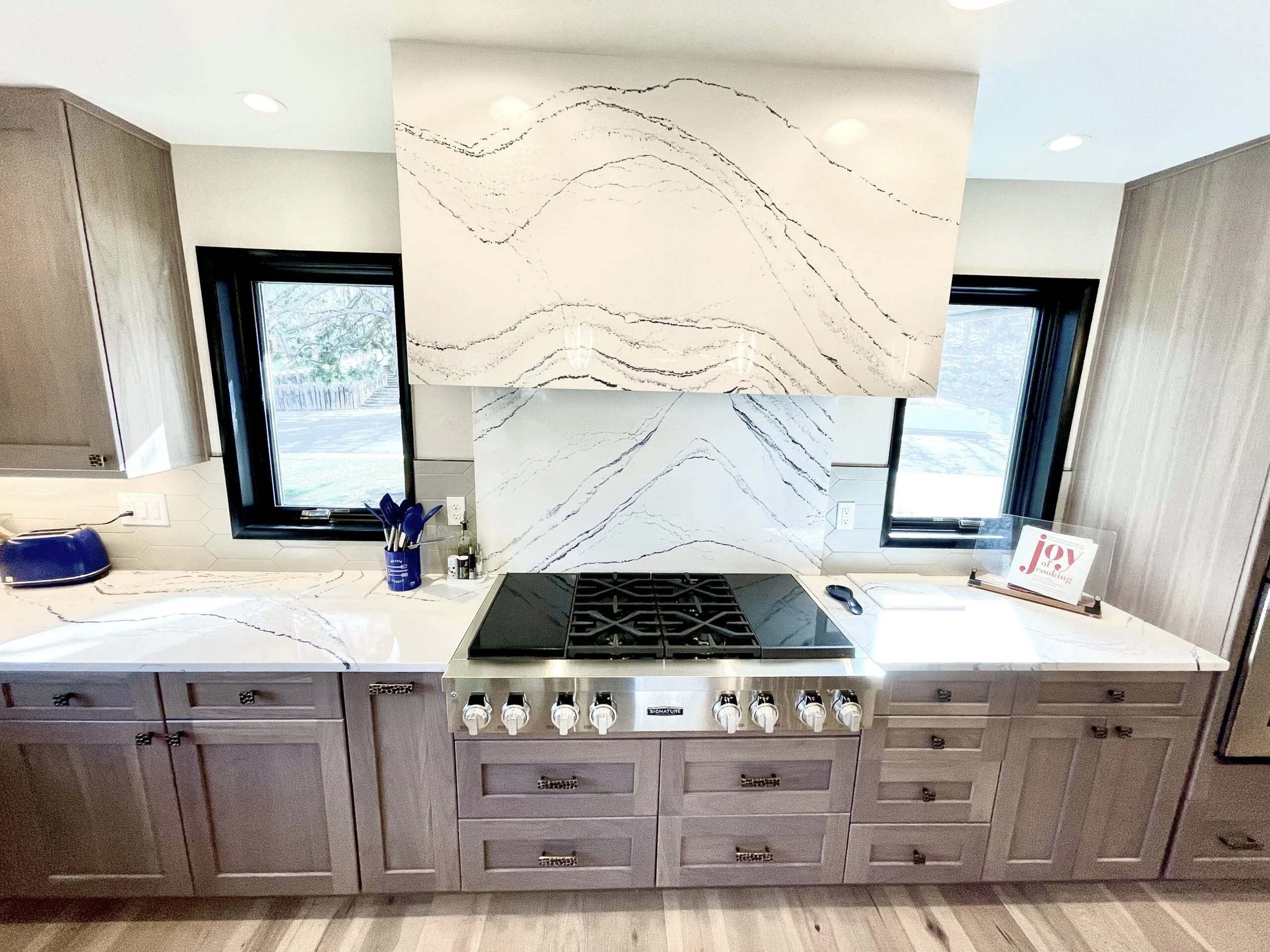 Kitchen with a marble backsplash, black window frames, and a gray wooden cabinet with drawers. A stove with control knobs is in the center. There is a small countertop with utensils and a greeting card that says "joy" on it.