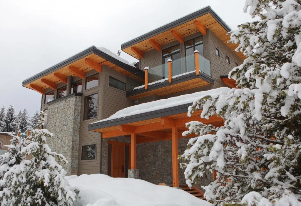 Modern house with large windows, wooden accents, stone details, and snow-covered trees and roof in a winter setting.
