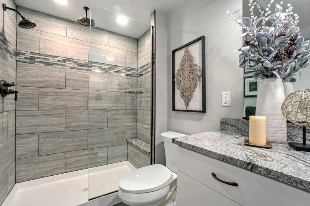 Bathroom with a tiled shower, a toilet, and a gray and black granite countertop with decorative items including a large vase with flowers, a candle, and a framed artwork on the wall.