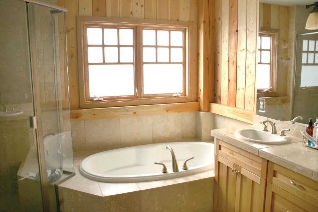 A bathroom with a large window above a built-in bathtub, wooden walls, a sink with a mirror, and a glass-enclosed shower.