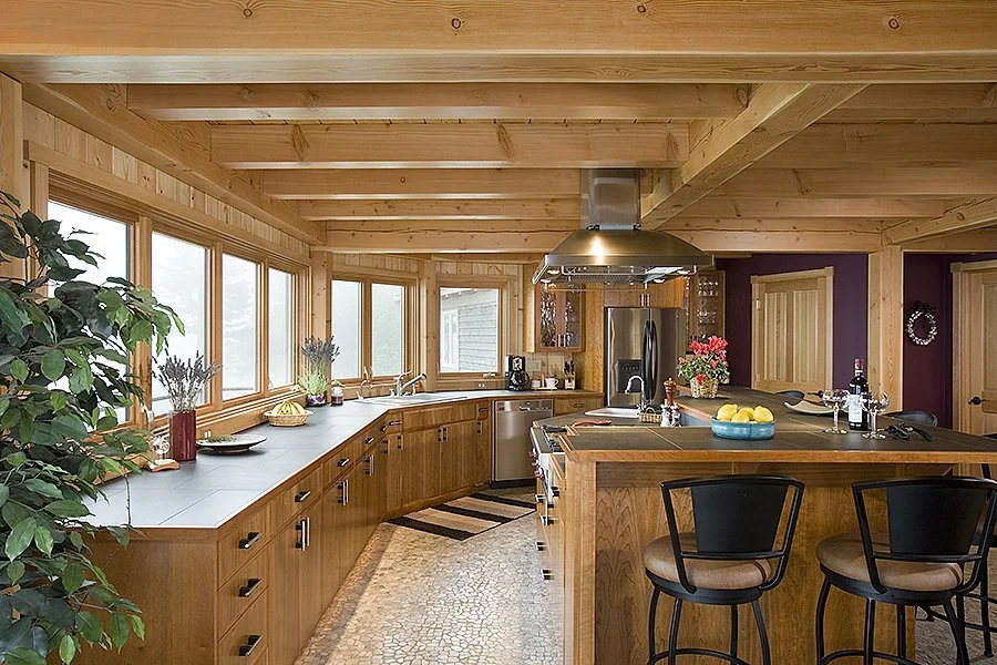 A wooden kitchen with natural wood cabinetry, large windows, and a curved countertop. There are various kitchen appliances, decorative items, and bar stools in the space.