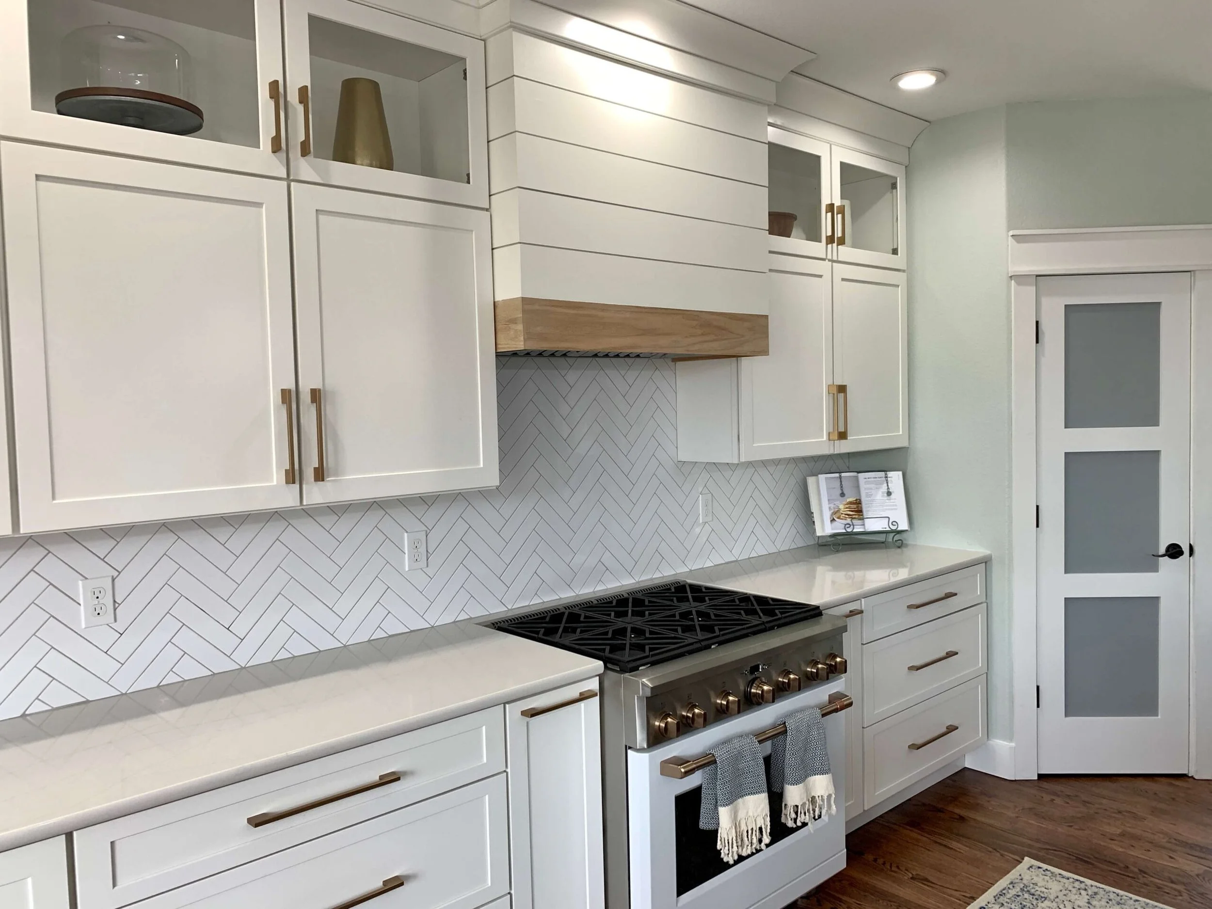 Modern kitchen with white cabinets, a marble countertop, a white herringbone backsplash, and a gold-tone stove with oven. Open upper cabinets display glassware and decorative items, and there are two towels hanging on the oven handle.