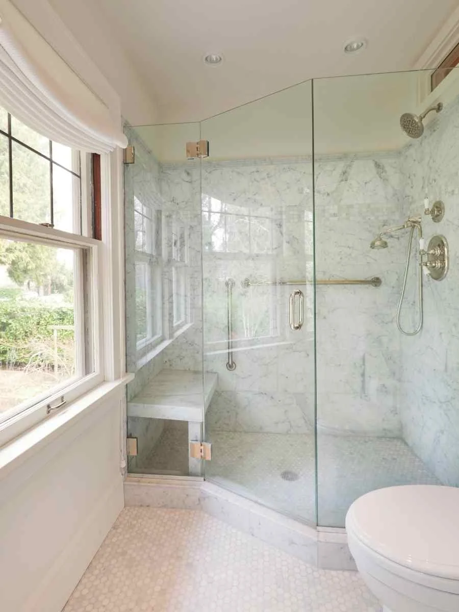 Bathroom with a glass-enclosed walk-in shower featuring marble walls and floor, a bench, two showerheads, a grab bar, a window with a view of greenery, and a white toilet.