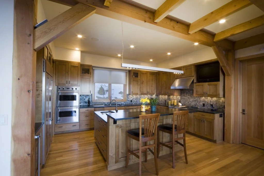 A wooden kitchen with a central island, stainless steel appliances, and a window above the sink. There are two chairs at the island, and a TV mounted on the wall.