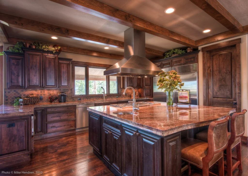 A spacious kitchen with dark wooden cabinets, a large granite island with seating, and stainless steel appliances. Wooden beams are on the ceiling, and there are large windows behind the sink with a view outside. A vase with flowers is on the island.