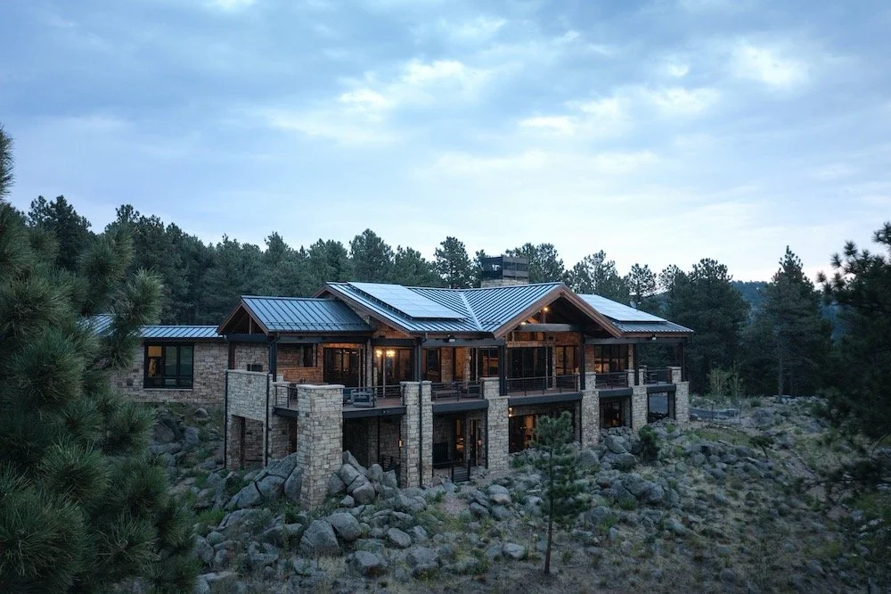 A modern house with stone and wood exterior, situated on a rocky hillside surrounded by trees, with a metal roof and illuminated windows.