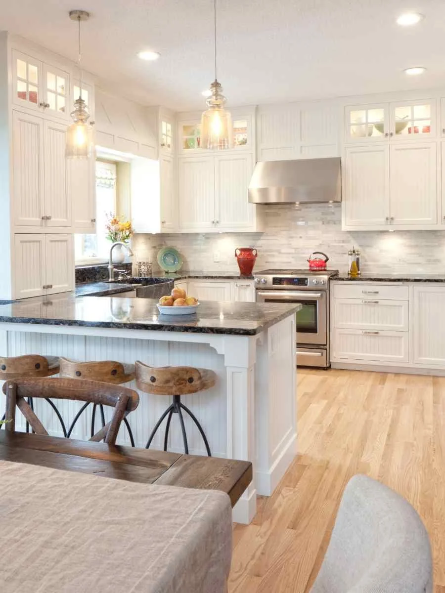 A bright kitchen with white cabinets, granite countertops, a stainless steel stove with a red teapot, a bowl of apples on the island, and wooden chairs at a dining table.
