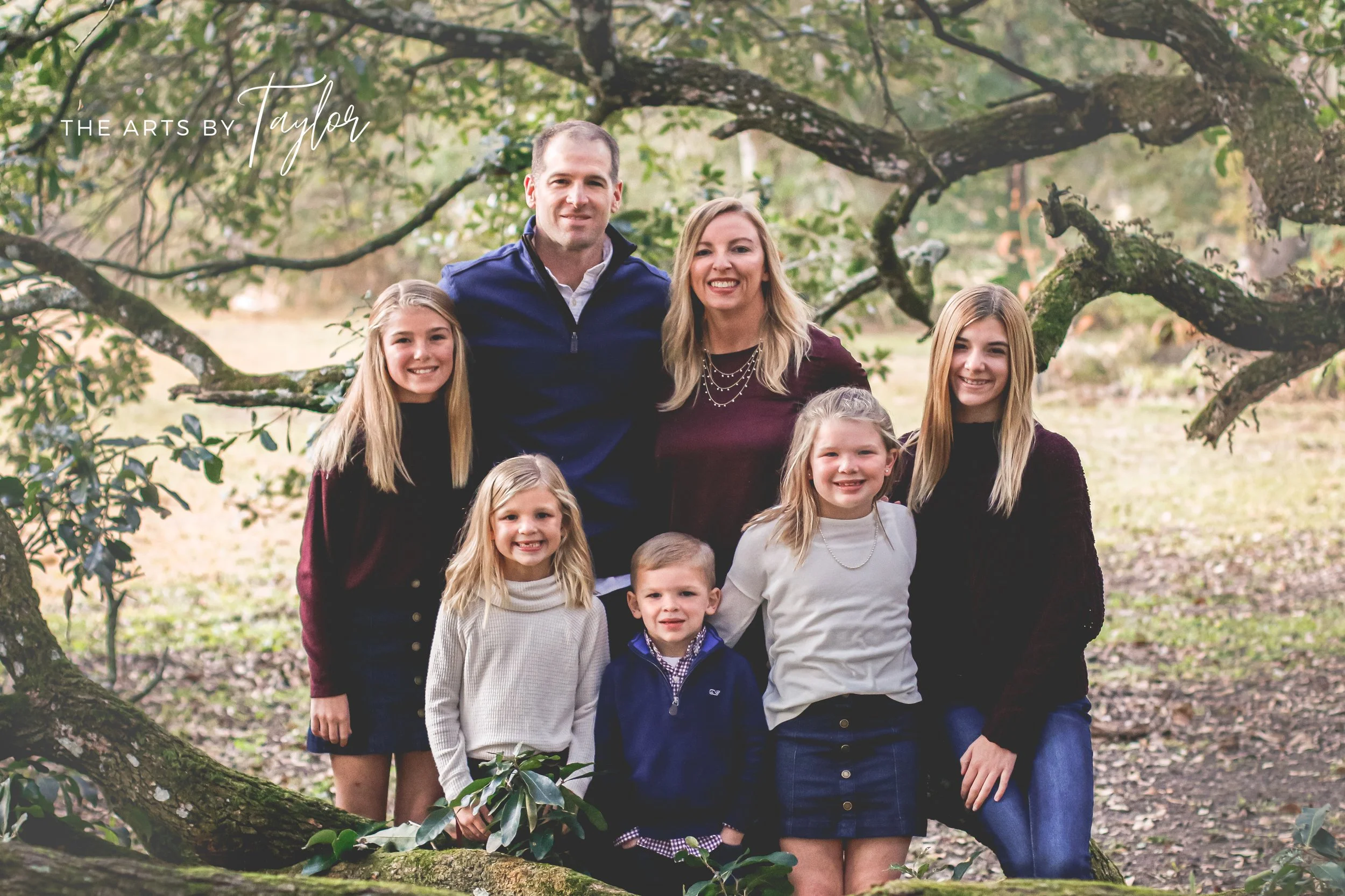 A family of seven people, including children and adults, standing outdoors on a large tree branch in a park or wooded area during daytime, smiling at the camera.