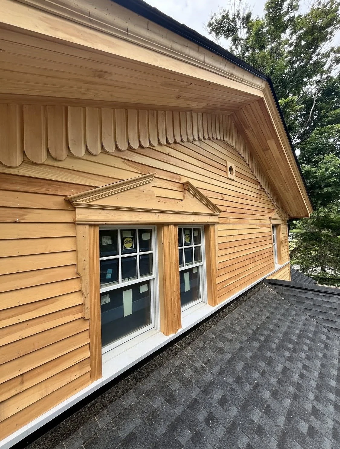 Close-up of a wooden house exterior showing detailed wood siding and double-pane windows with decorative pediments, set against a background of trees.
