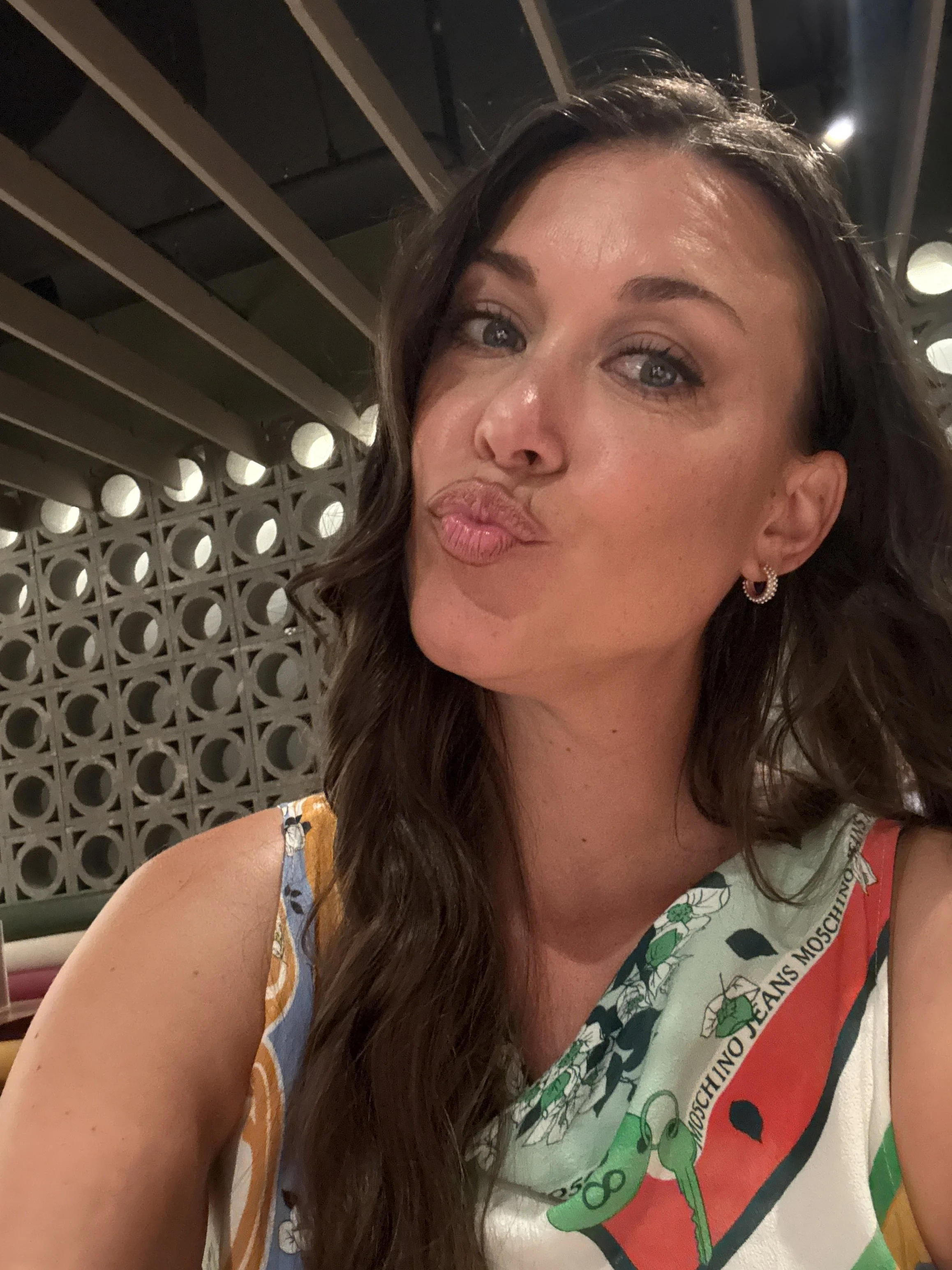 A woman making a kissy face with pursed lips, wearing earrings and a colorful shirt with a logo that includes a green character and text, in a setting with a decorative wall and ceiling in the background.