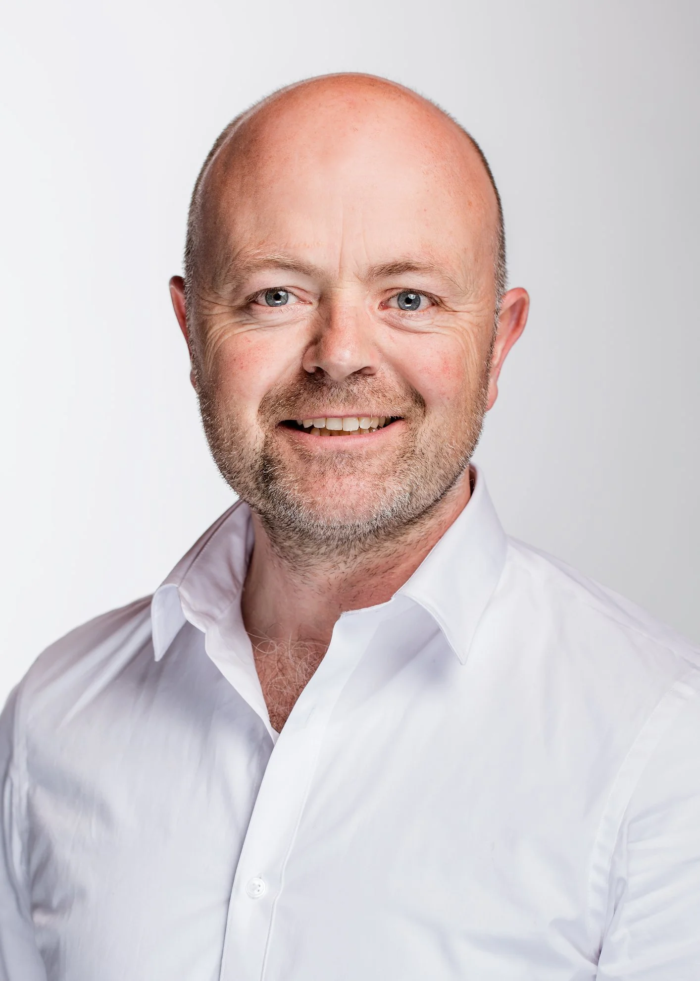 Portret van een man met een glimlach, een baard en een kale kop, gekleed in een witte blouse, tegen een egale lichte achtergrond.