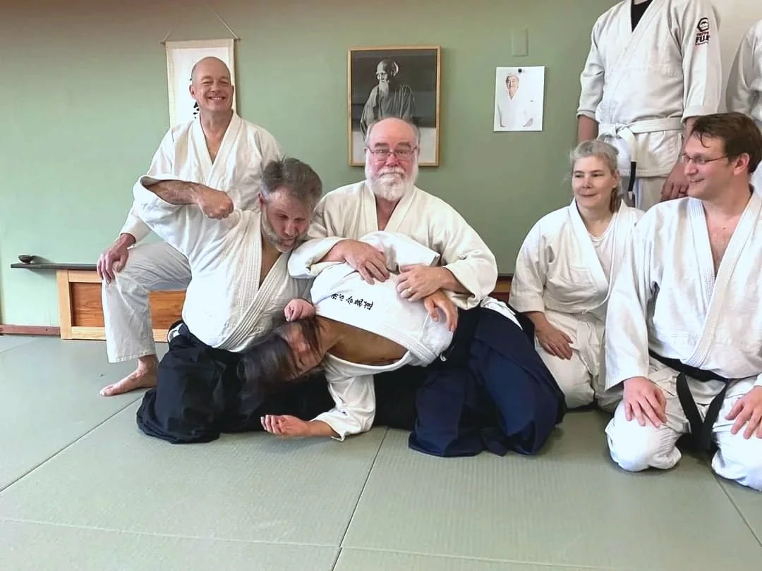 Group photo with instructors James Baker Shihan, Steve Nelson, and Eric Pierce with students in front of the Aikido of Norfolk's kamiza and photo of Morihei Ueshiba the founder of Aikido.