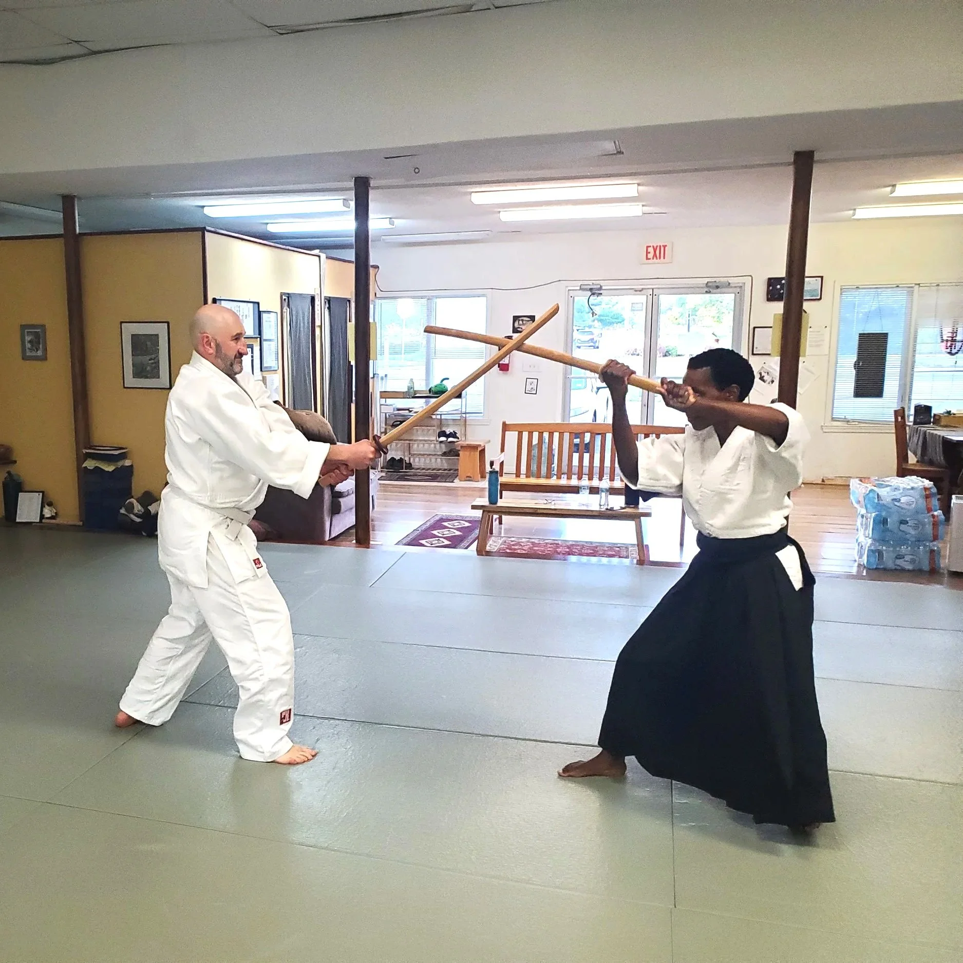 Two martial artists practicing martial arts with wooden bokens in the dojo at Aikido of Norfolk. One person is wearing a white gi, and the other is in a black hakama and a white top.