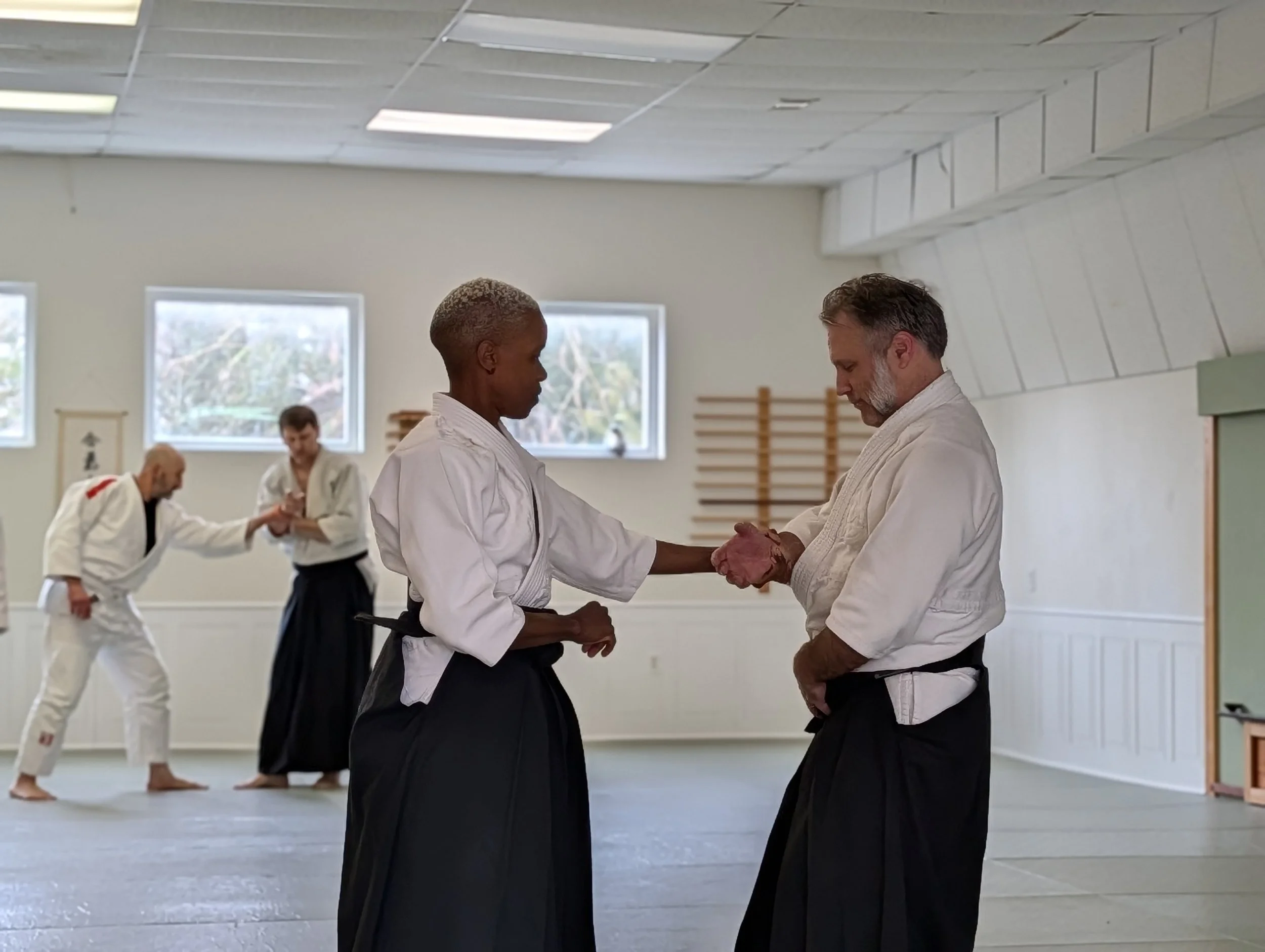 Two martial artists with white gis and hakamas practicing Aikido, at Aikido of Norfolk. Uke is attacking nage with katatetori in a dojo with other students in the background.