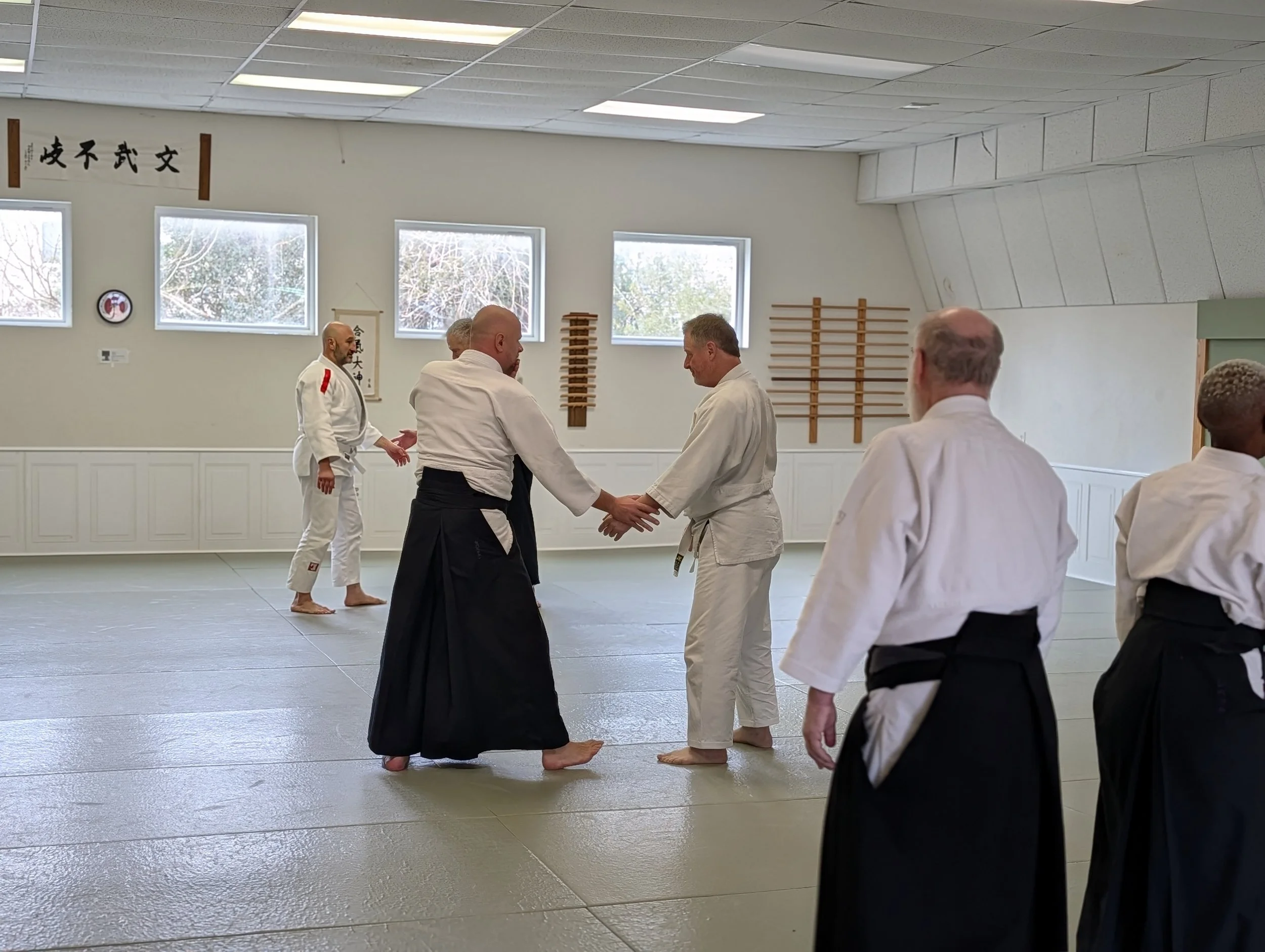 Martial arts class, at Aikido of Norfolk, with students in uniforms practicing Aikido on a padded mat, with instructor James Baker Shihan supervising.