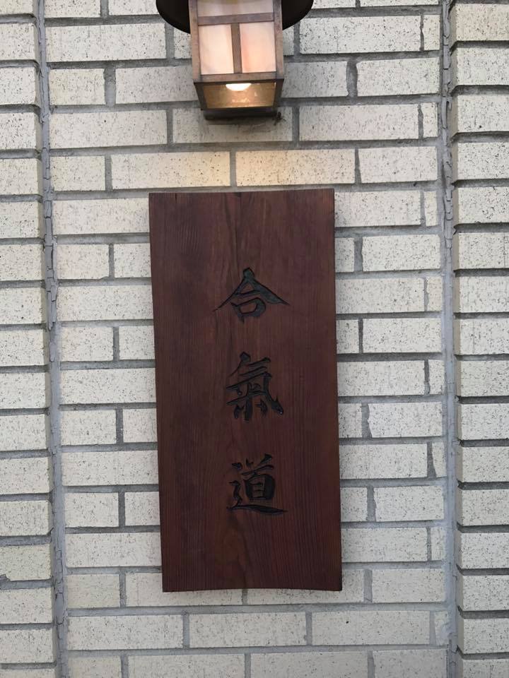 Wooden sign with Japanese characters stating "Aikido" mounted on the brick wall of Aikido of Norfolks building, illuminated by an exterior light fixture above.