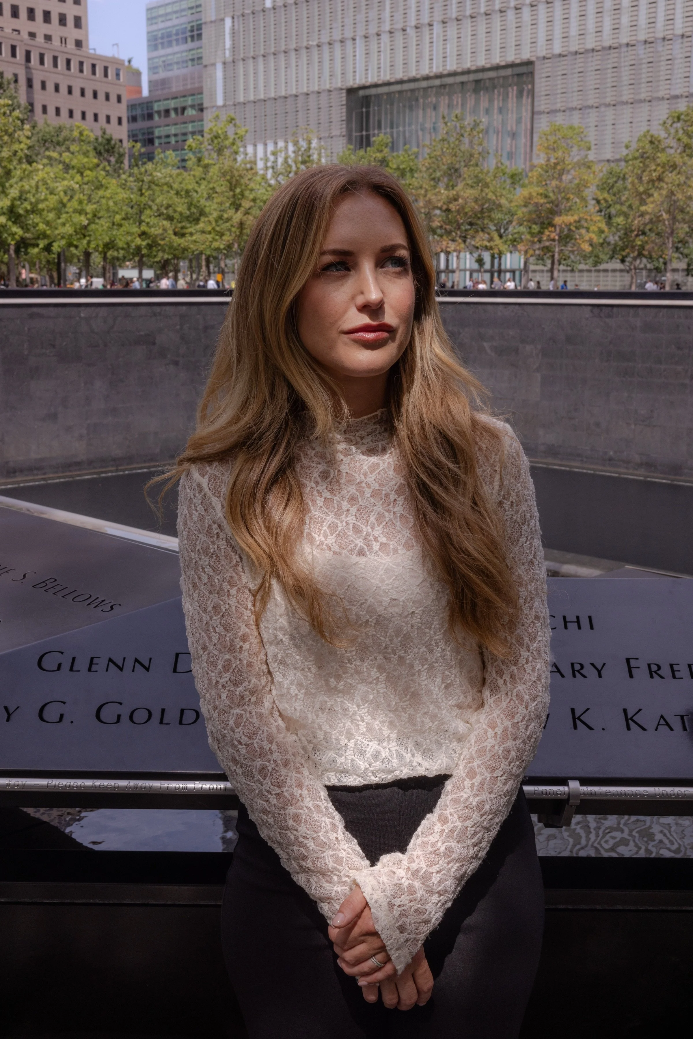 Ashley Bisman with long, wavy, light brown hair standing in front of the 9/11 memorial with names inscribed on it, in downtown New York City.