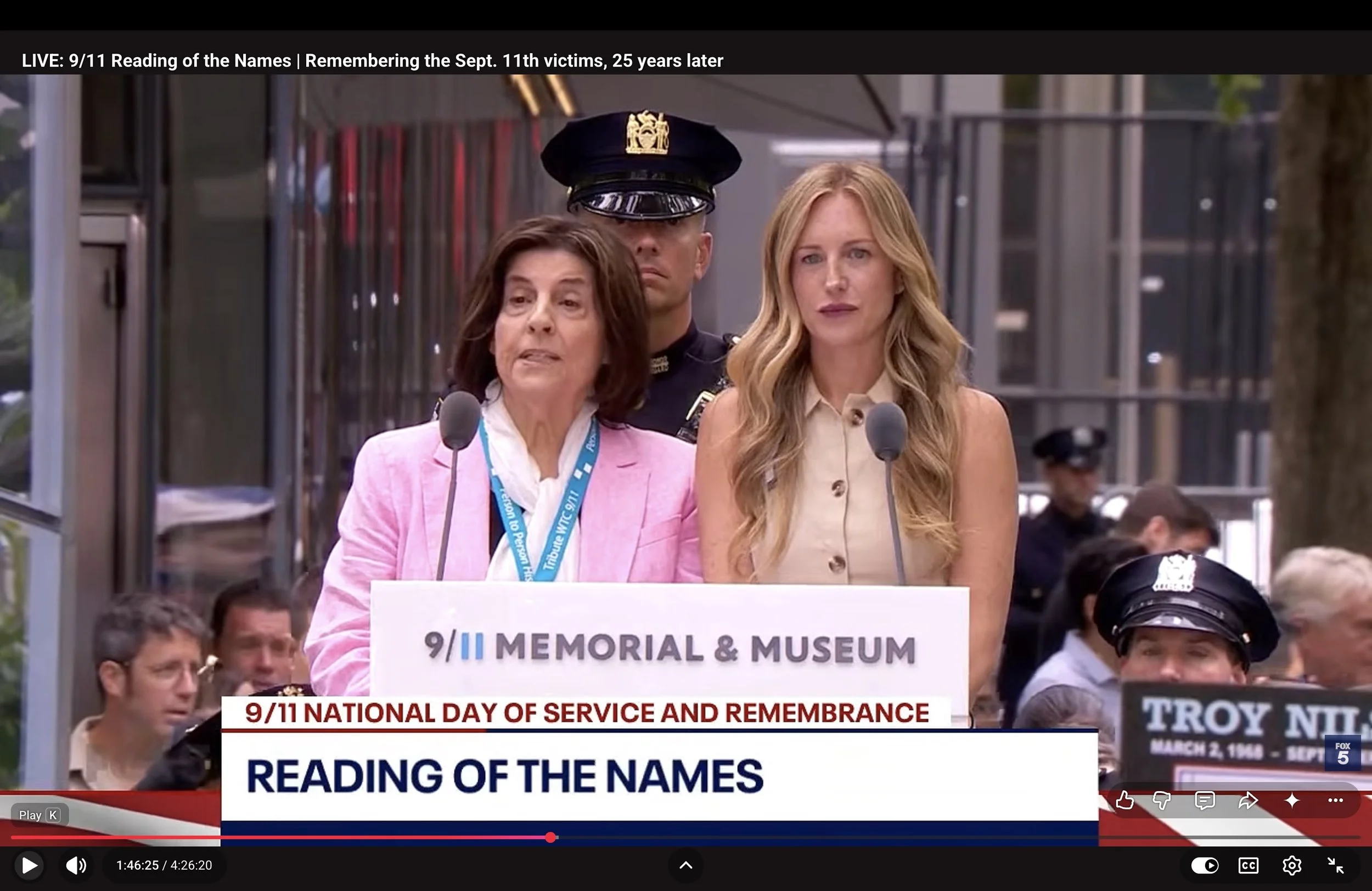A woman in a pink blazer speaking at a podium during a memorial event, with a police officer behind her, in front of a crowd. The event is related to 9/11 remembrance.