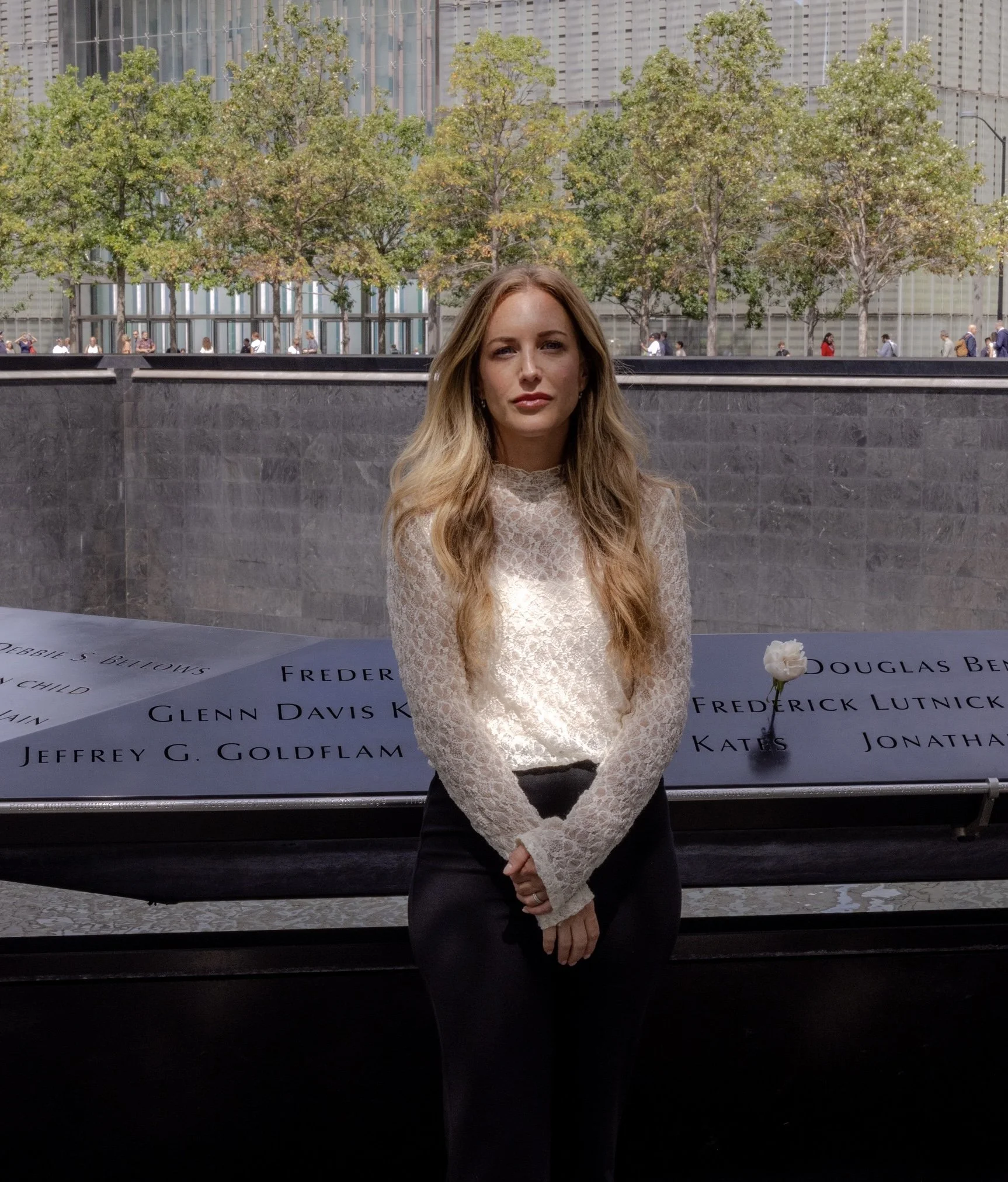 A woman with long blonde hair wearing a white lace top and black pants, standing in front of a memorial with names inscribed on it, and trees and a city building in the background.