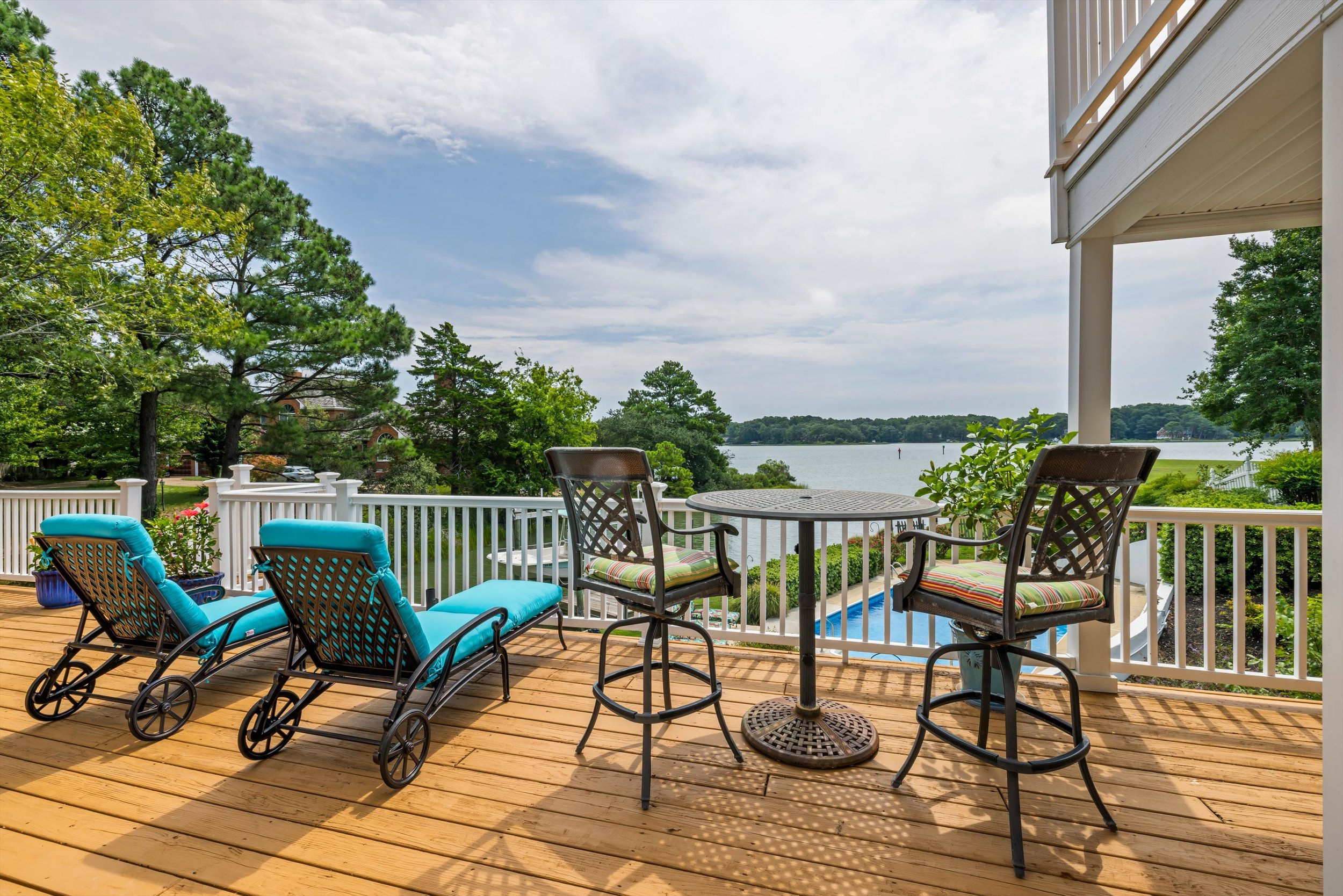 A wooden deck with outdoor furniture, including a round table, chairs with cushions, and a lounge chair. The deck overlooks a lake with trees and a partly cloudy sky in the background.