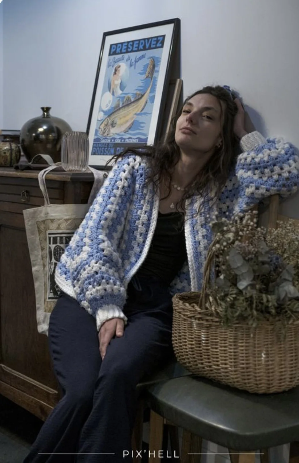Une jeune femme assise contre un mur avec une main dans ses cheveux, portant un pull en crochet bleu et blanc, à côté d'un panier de fleurs séchées. Un tableau avec une mer, un poisson et le mot 'PREservez' est posé derrière elle.