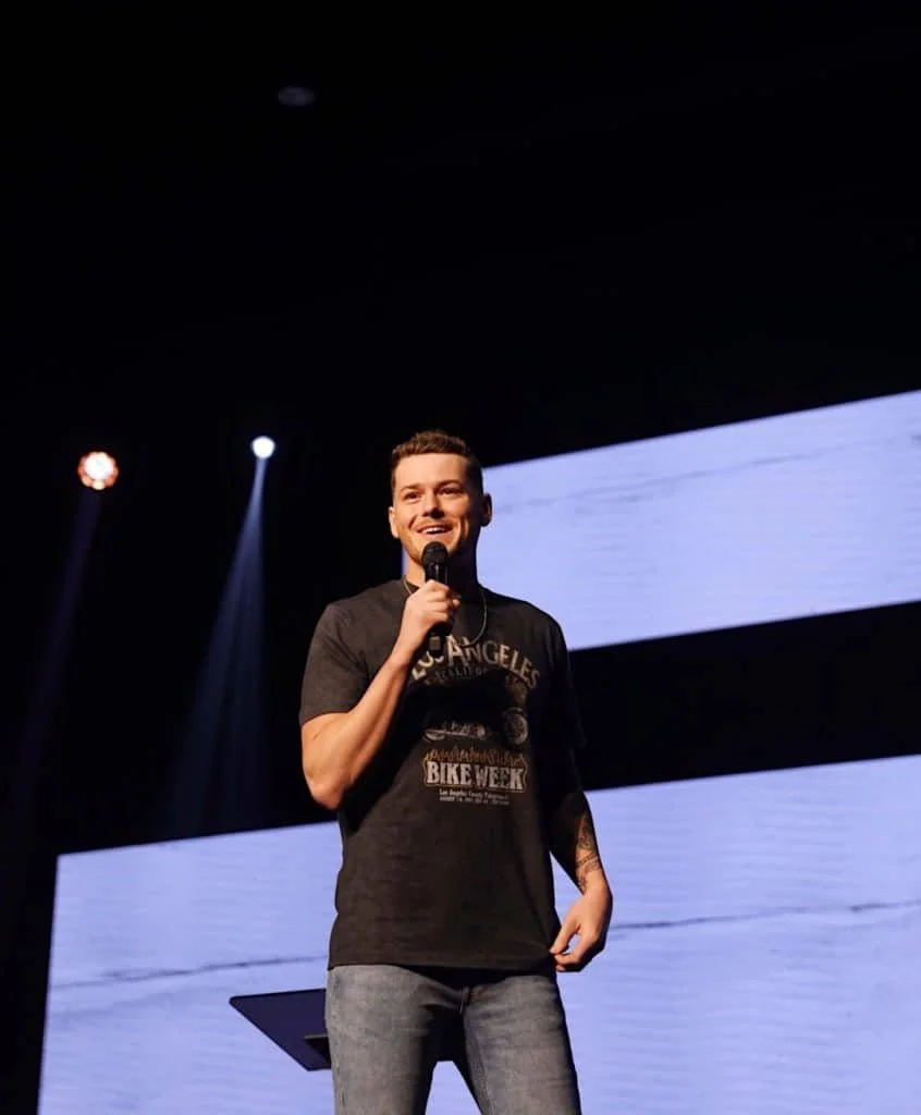 Young man smiling and speaking into a microphone on stage, wearing a black T-shirt and jeans.